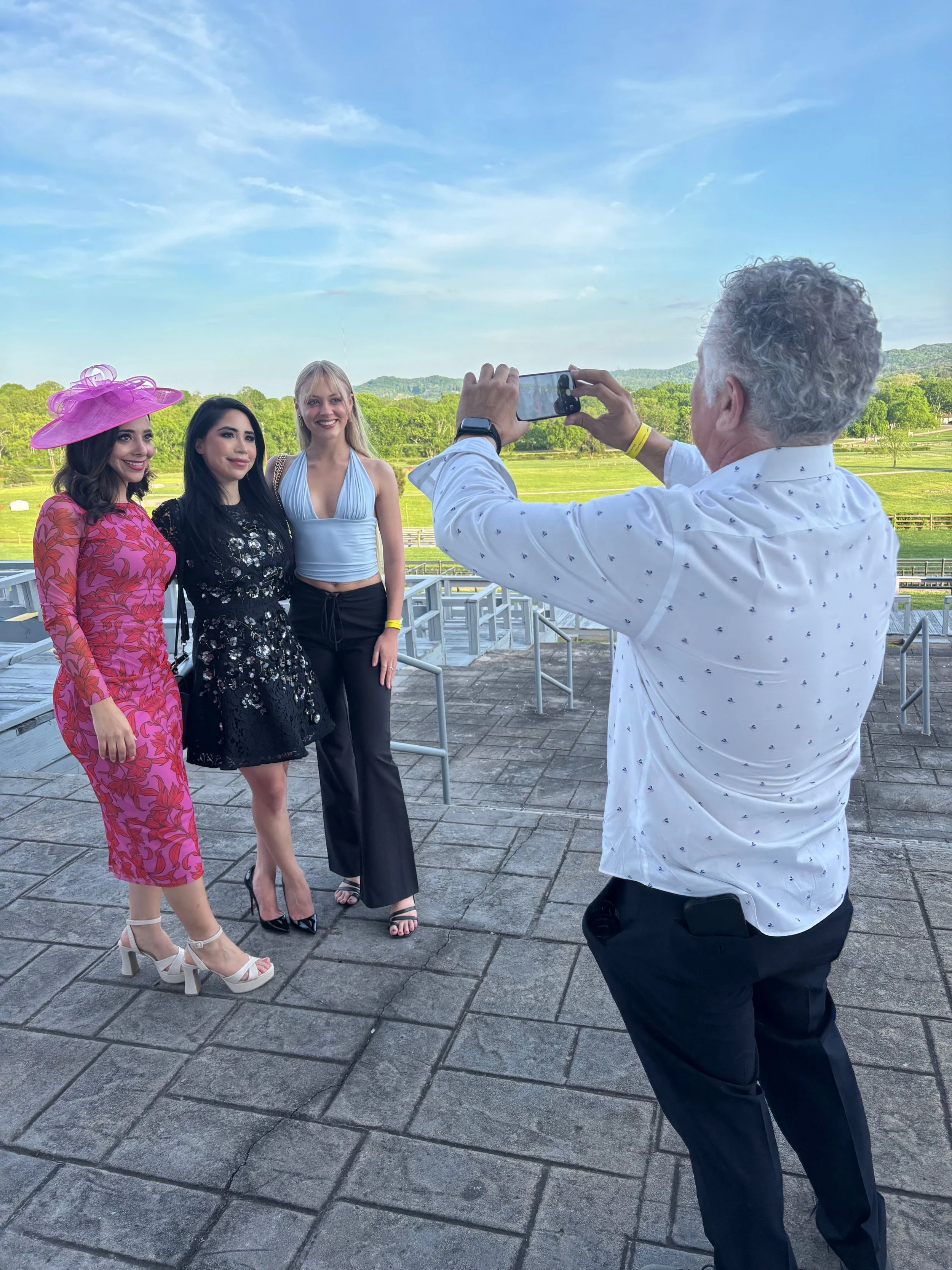 Three women taking a photo with a man at the racetrack area outdoors, with green fields and trees in the background.