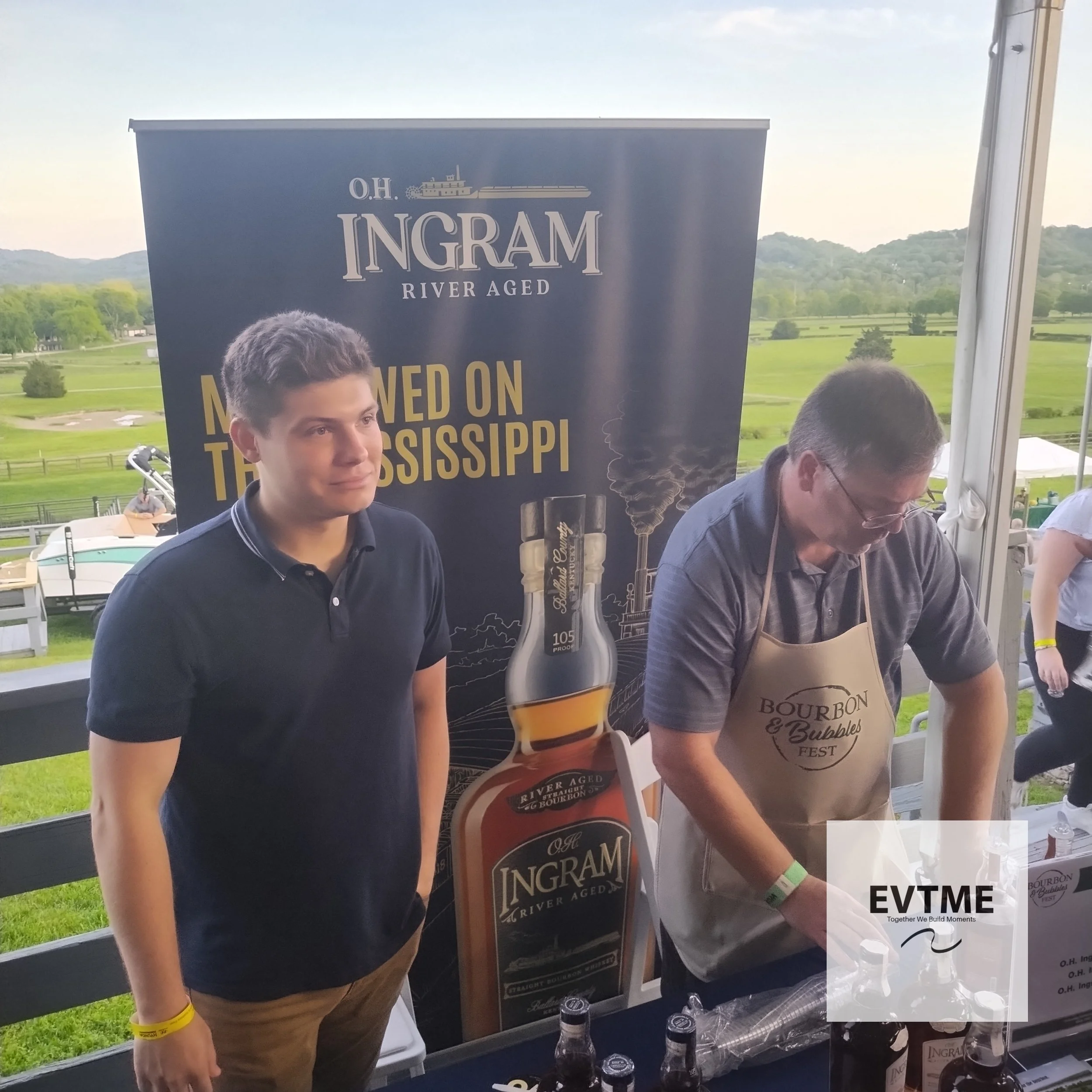 Two men at a bourbon tasting event, with a backdrop advertising O.H. Ingram River Aged bourbon and a bottle display, during daytime at an outdoor venue with green fields and mountains in the background.
