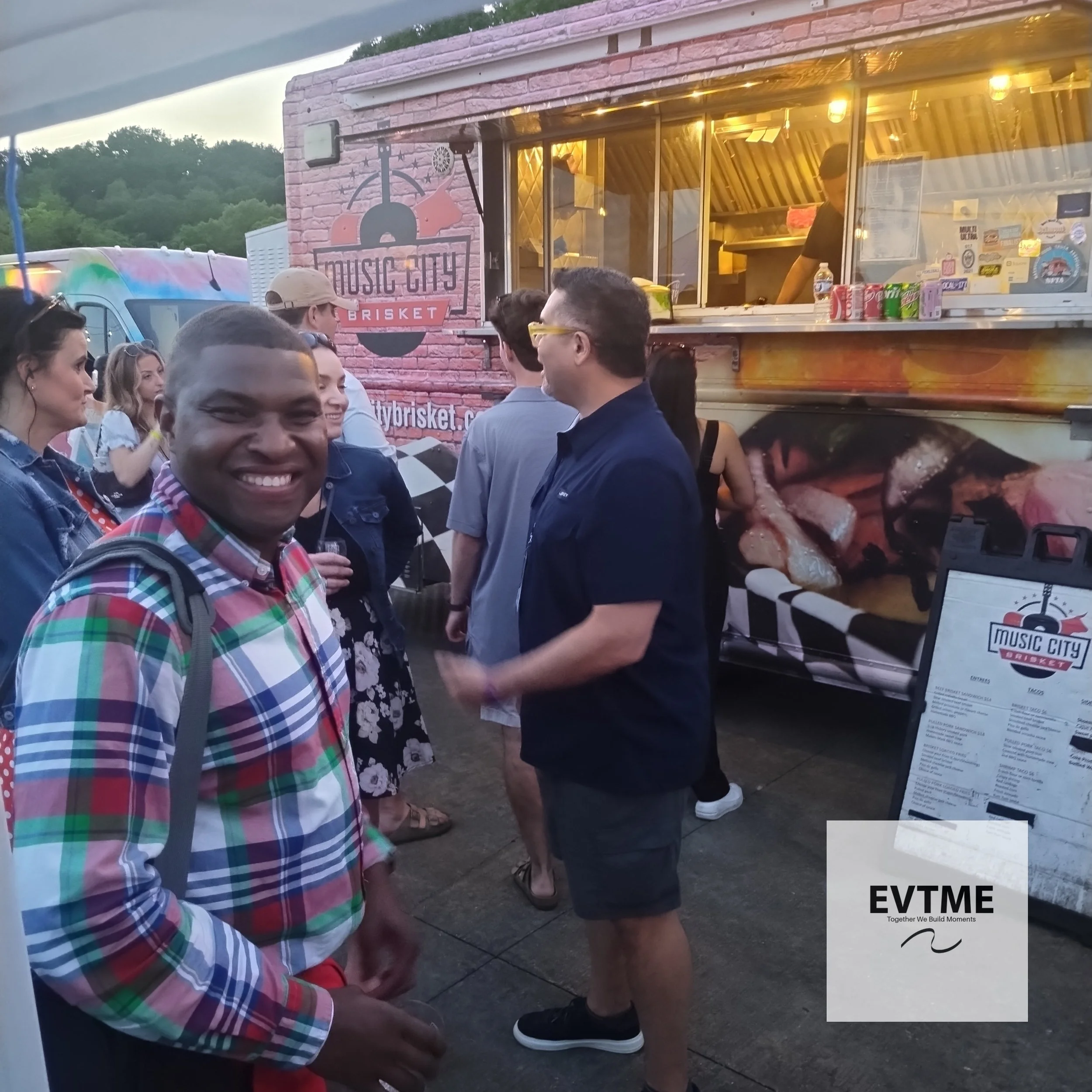 A group of people standing in line at a food truck called 'Music City Brisket' during an outdoor event at dusk. One man in the foreground is smiling at the camera, wearing a colorful plaid shirt and carrying a backpack.
