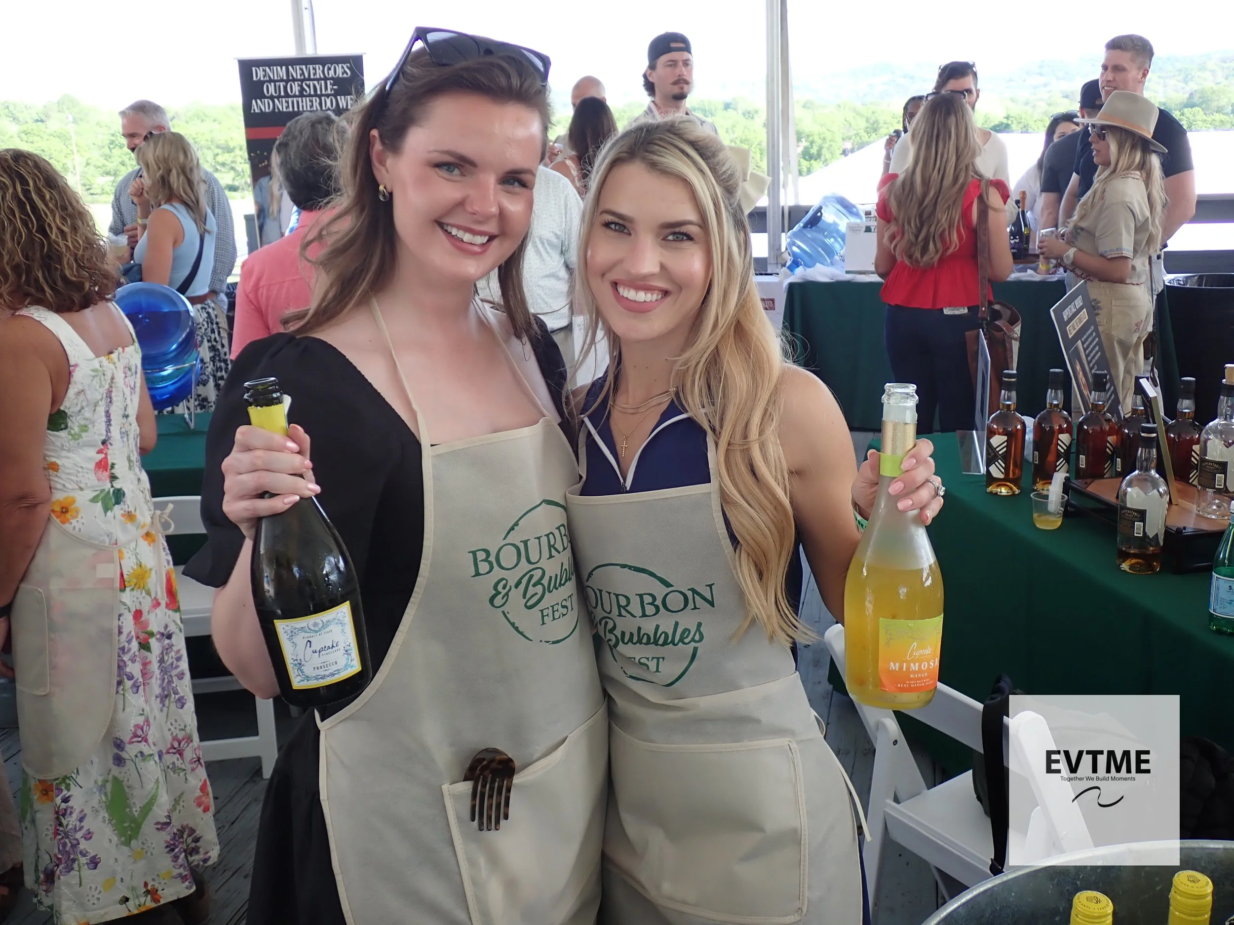 Two women wearing aprons at an outdoor bourbon and bubbles festival, holding bottles of drinks, with other festival attendees and booths in the background.