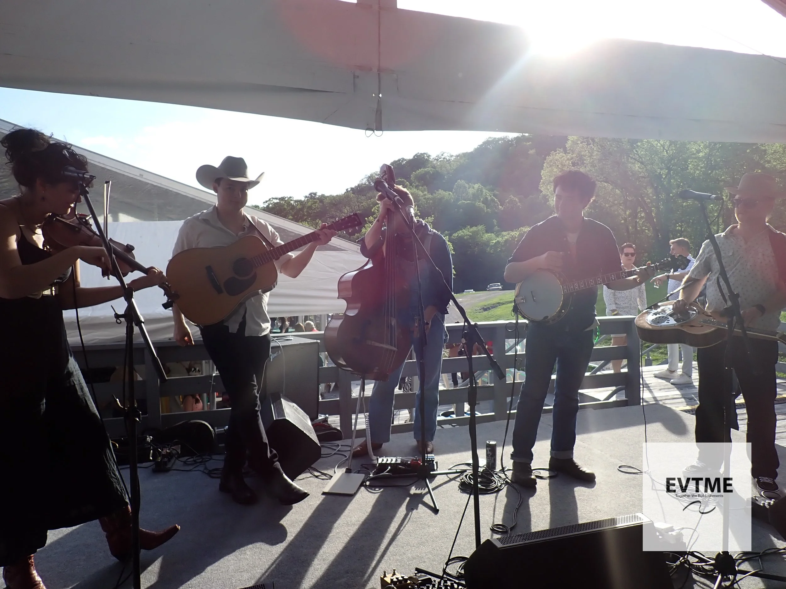 A group of musicians performing outdoors on a stage, playing guitars and a violin, with trees and sunlight in the background.