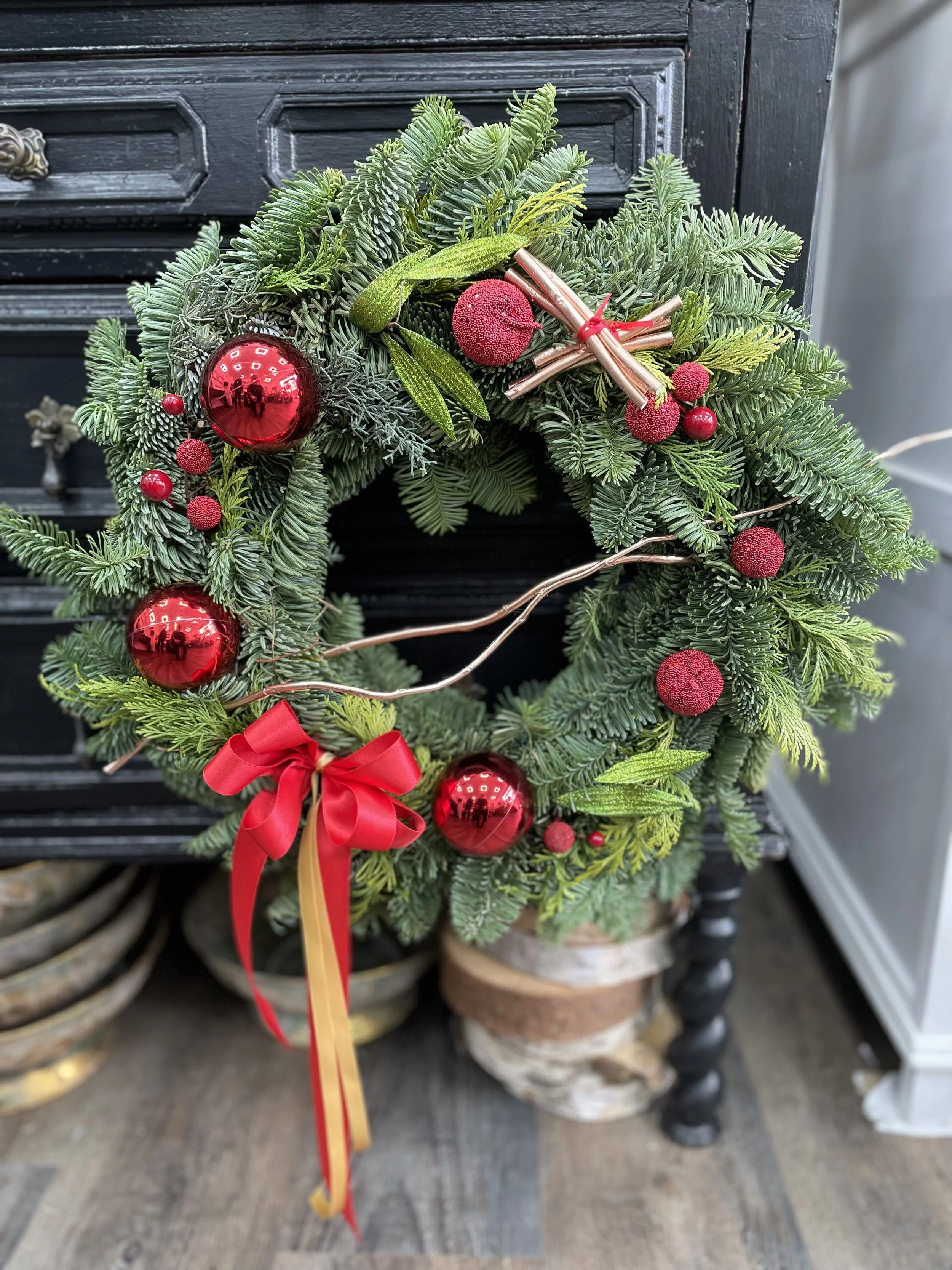 Christmas wreath with red ornaments, a red bow, and greenery including pine and fir branches, hanging on a black piece of furniture.