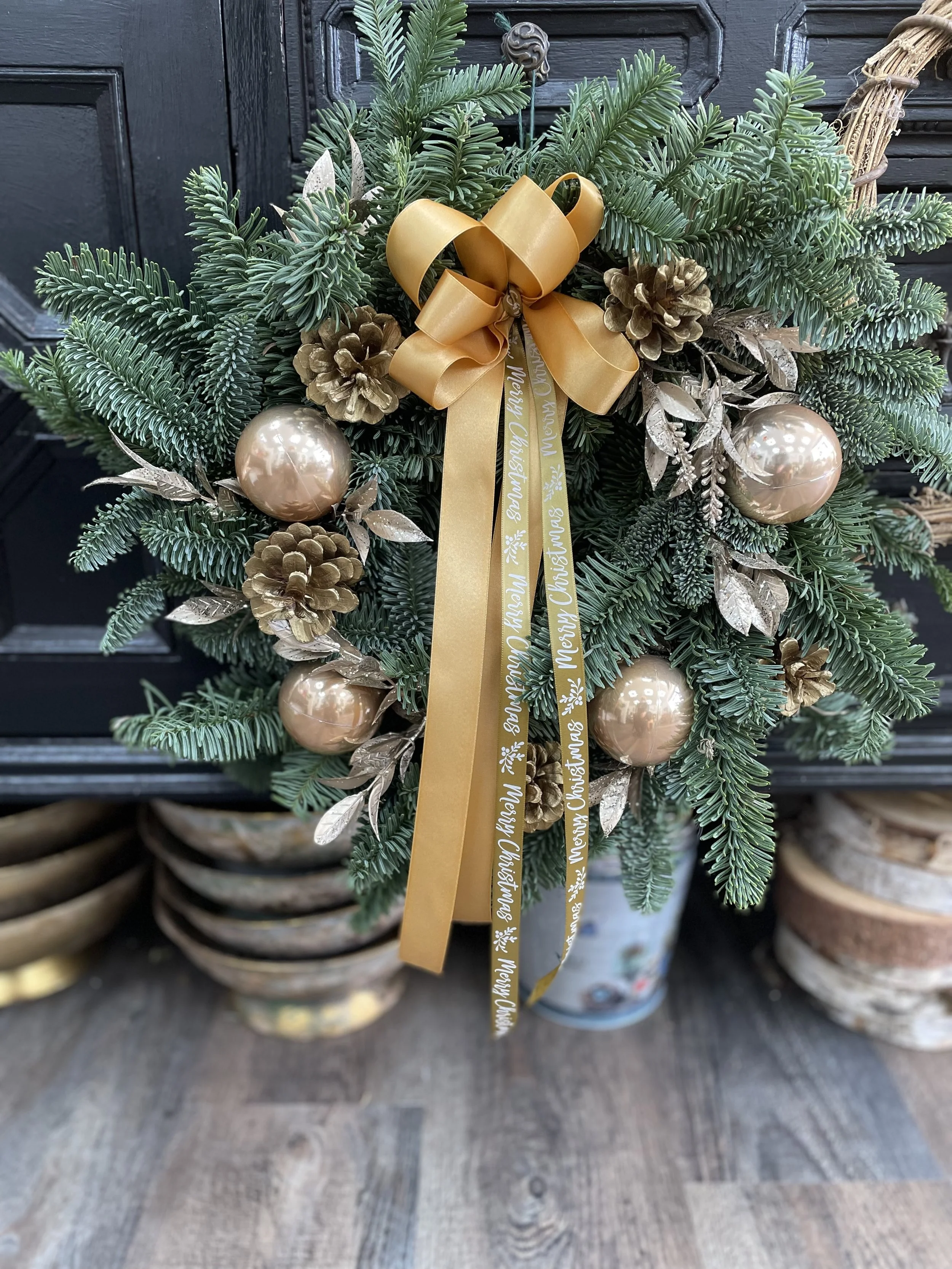 Christmas wreath made of evergreen branches decorated with gold ornaments, pinecones, painted leaves, and a gold ribbon with holiday messages, displayed in a pot on a wooden surface.