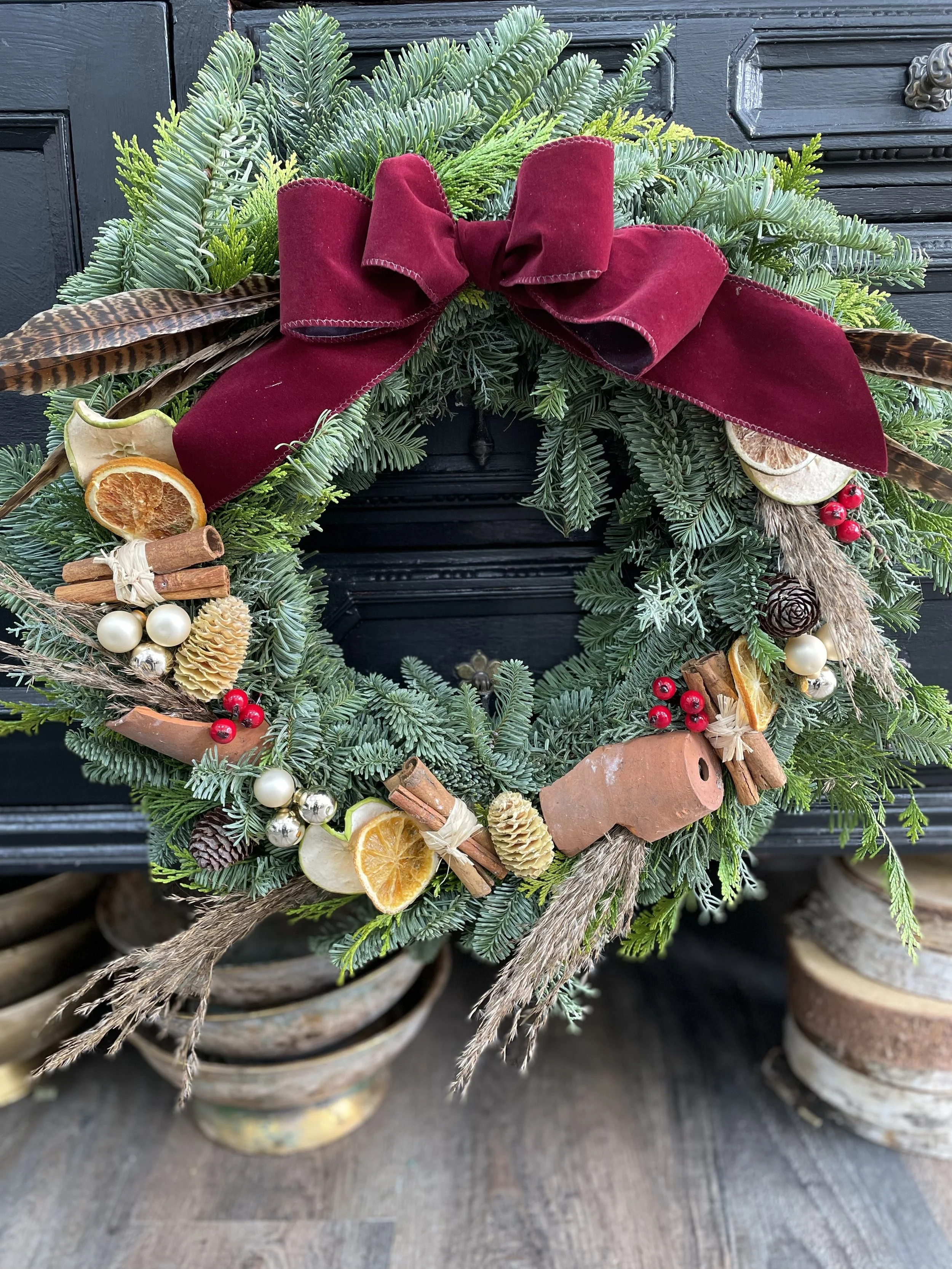 A Christmas wreath decorated with pine branches, a large burgundy bow, dried orange slices, cinnamon sticks, pine cones, red berries, white and silver ornaments, and dried flowers.