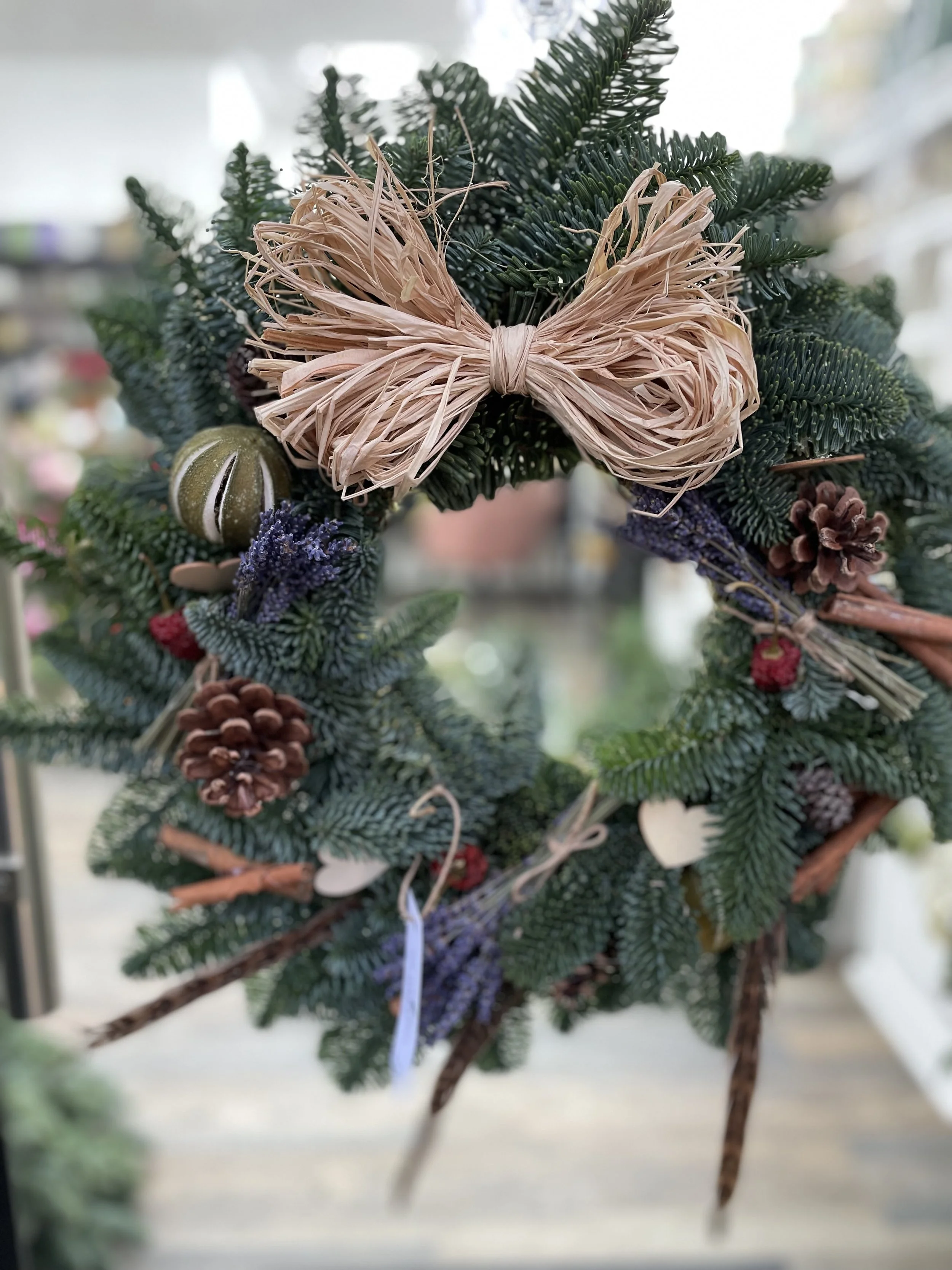 Close-up of a Christmas wreath decorated with pine branches, pinecones, small ornaments, and a beige bow made of straw.