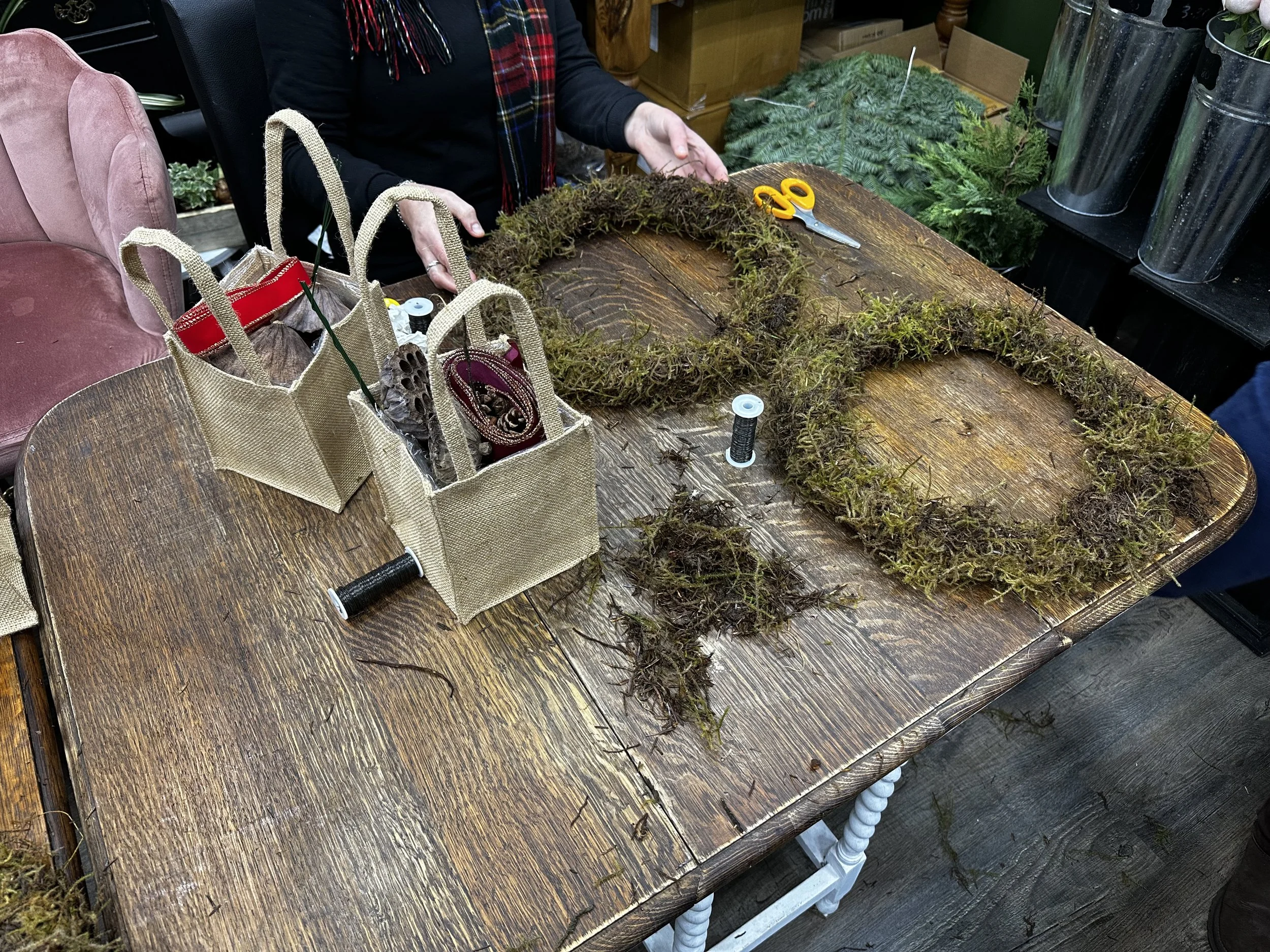 Person creating a Christmas wreath with moss and natural materials on a wooden table, with scissors, thread, and craft supplies nearby.