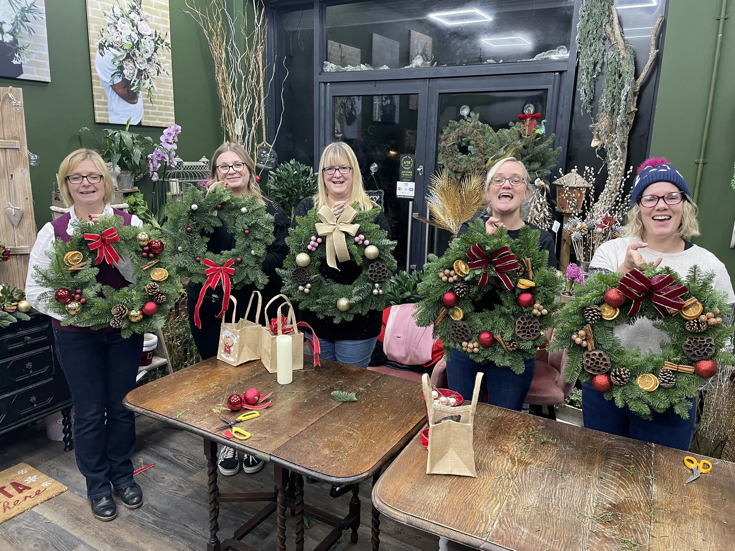 Five women standing behind a table, each holding a Christmas wreath decorated with ornaments, ribbons, and natural elements, inside a decorated craft store.