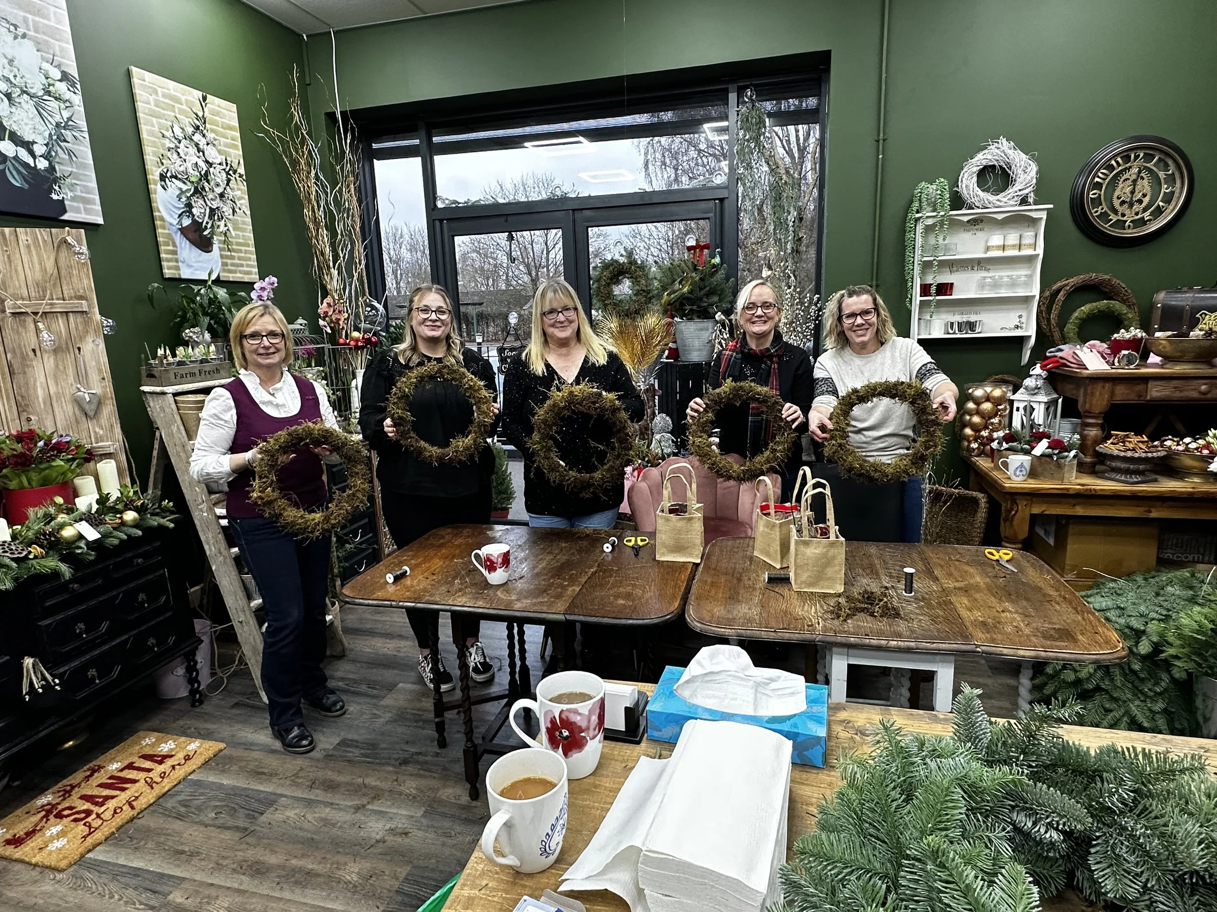 Five women standing behind a table in a decorated room, each holding a Christmas wreath they made. The room features Christmas decorations, gift bags, and crafting supplies on the table and surrounding shelves.