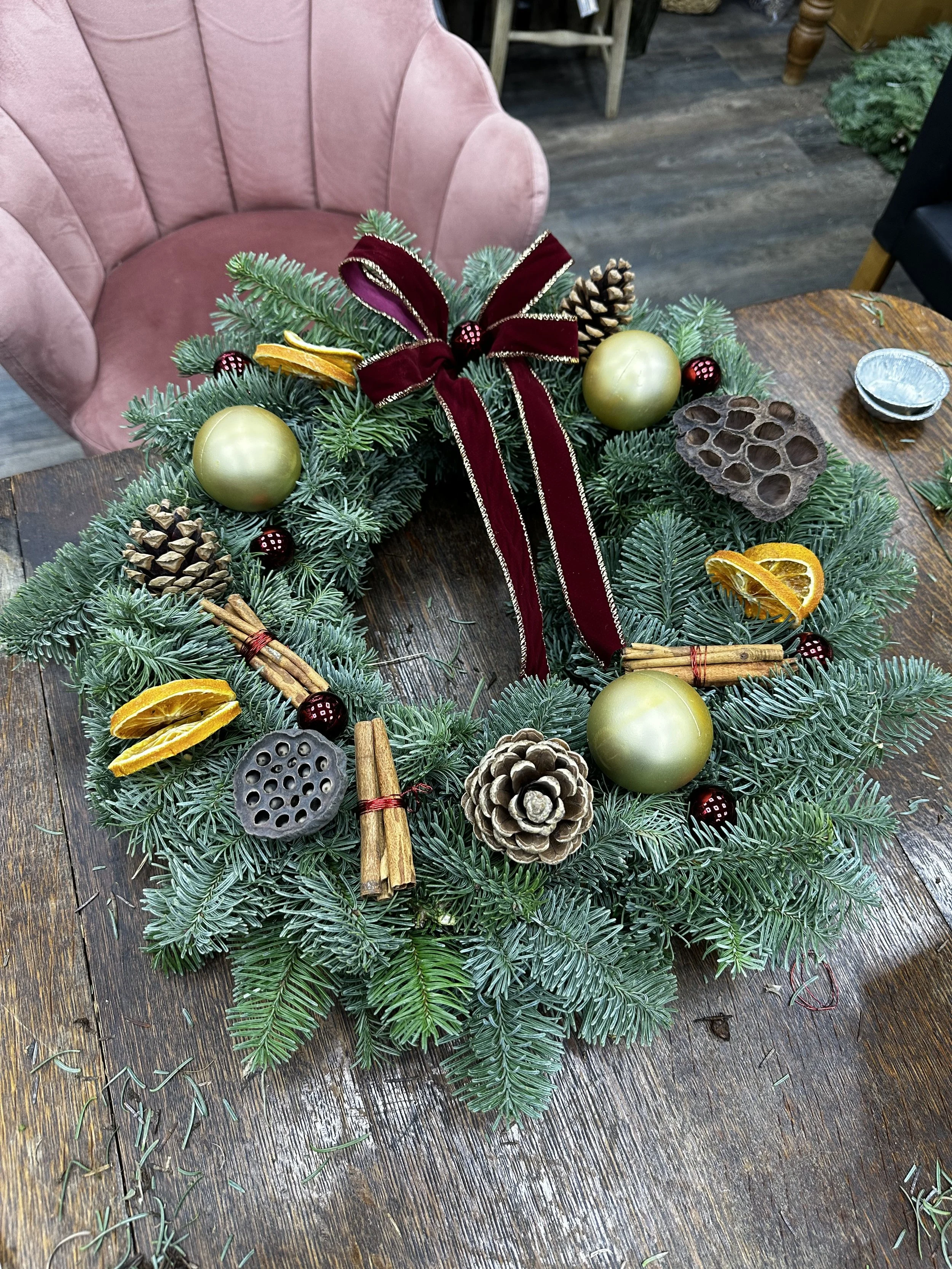 Christmas wreath with pine branches, gold ornaments, dried orange slices, cinnamon sticks, pinecones, a lotus seed pod, and a burgundy ribbon bow on a wooden table.