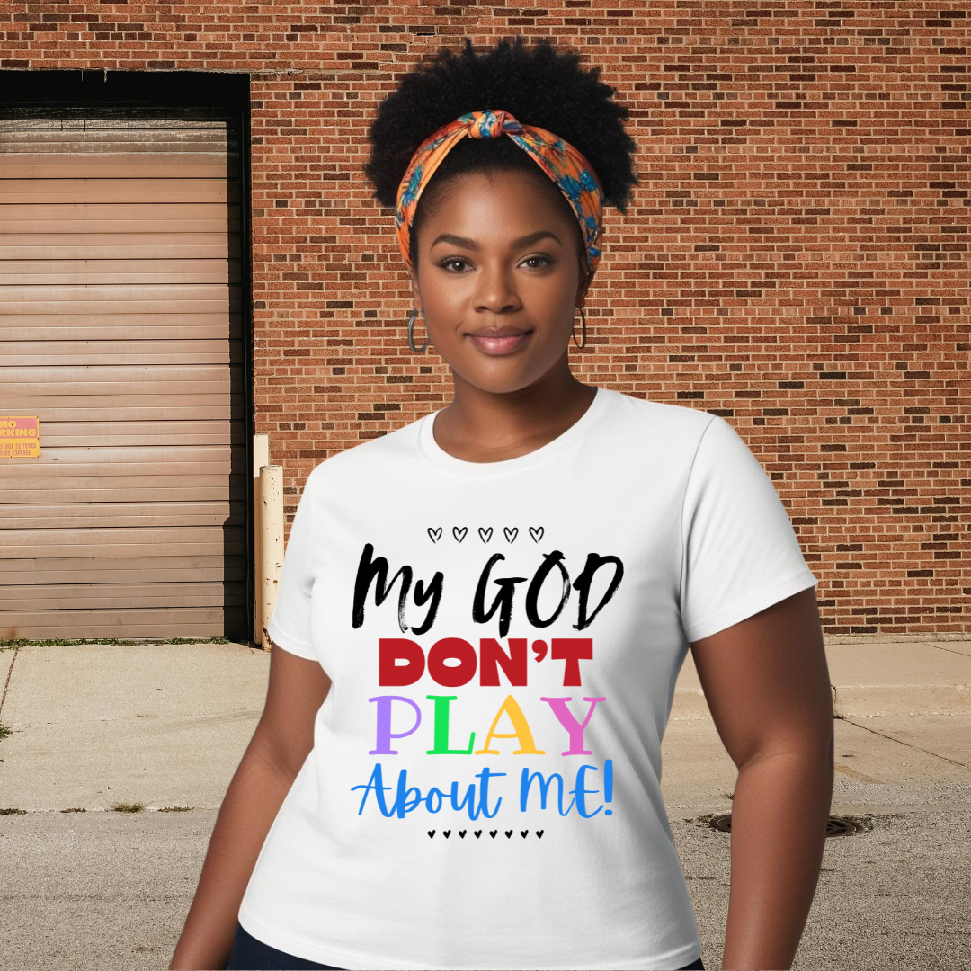 A woman standing outdoors in front of a brick wall, wearing a white T-shirt with colorful text that reads, "My GOD DON'T PLAY About ME!" She has a colorful headscarf, hoop earrings, and a confident expression.