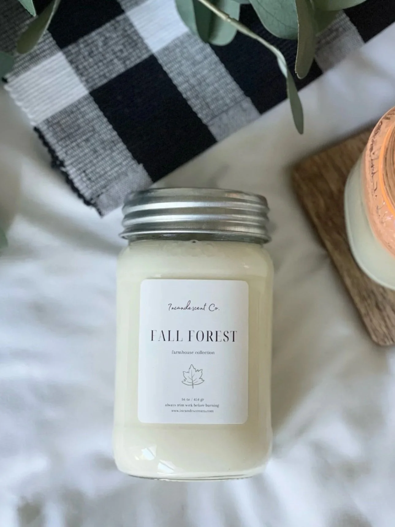 A candle labeled 'Fall Forest' from the Incandescent Co. farmhouse collection, placed on a white surface with a black and white checkered cloth and a plant in the background.