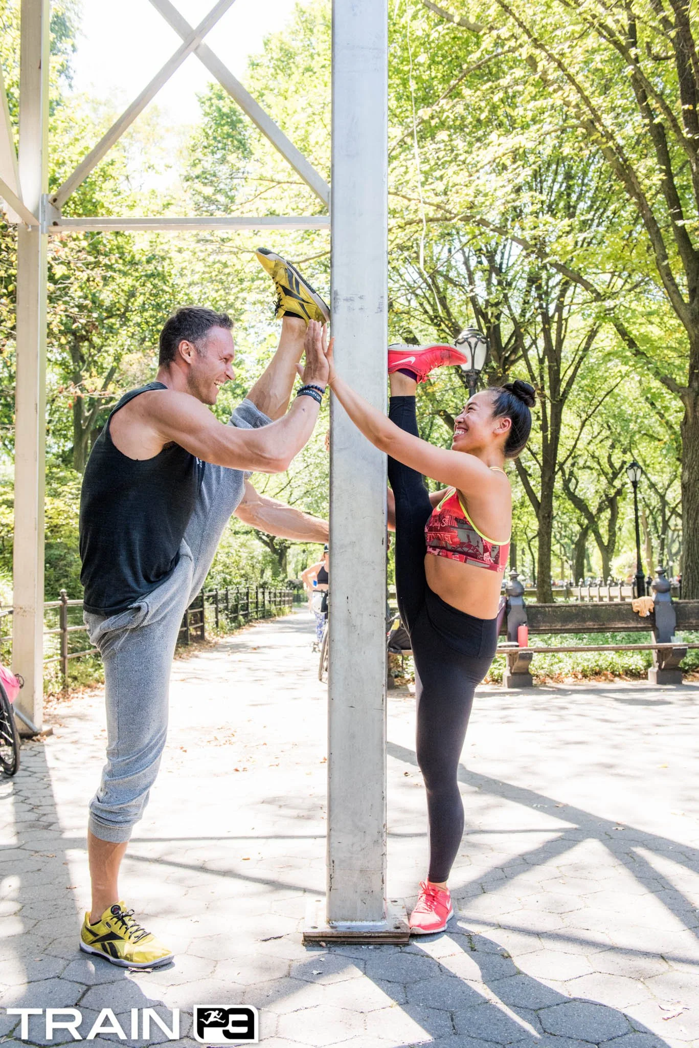 A man and woman practicing a fitness routine outdoors in a park, standing on either side of a vertical metal pole, touching palms, smiling, with trees and sunlight in the background.