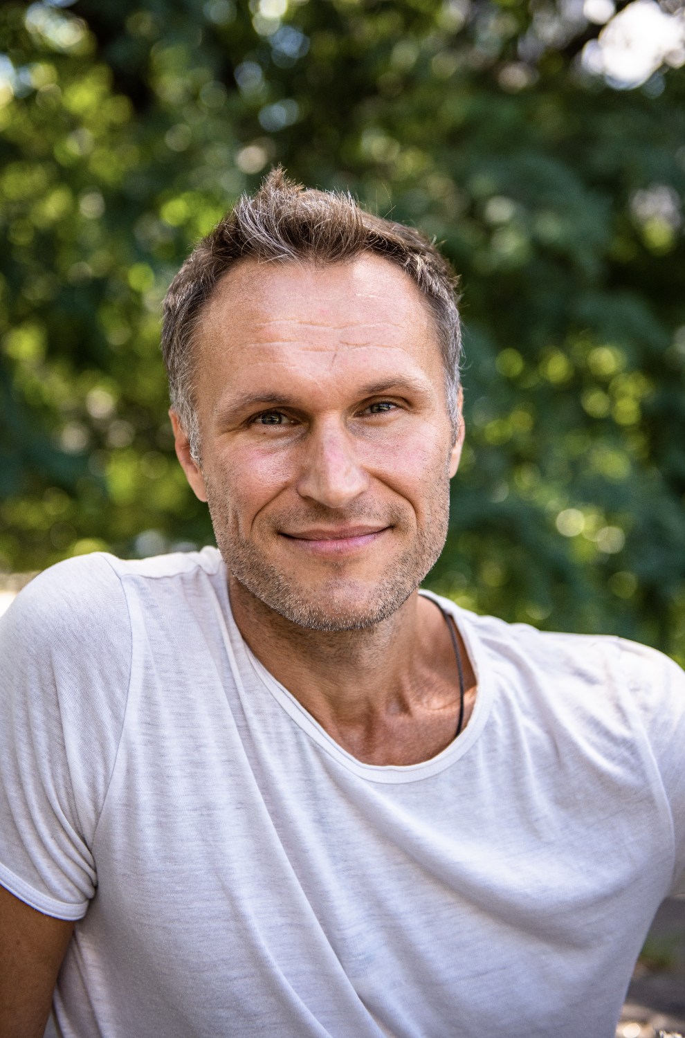 A smiling middle-aged man with short brown hair and stubble, wearing a white t-shirt outdoors with a background of green trees.