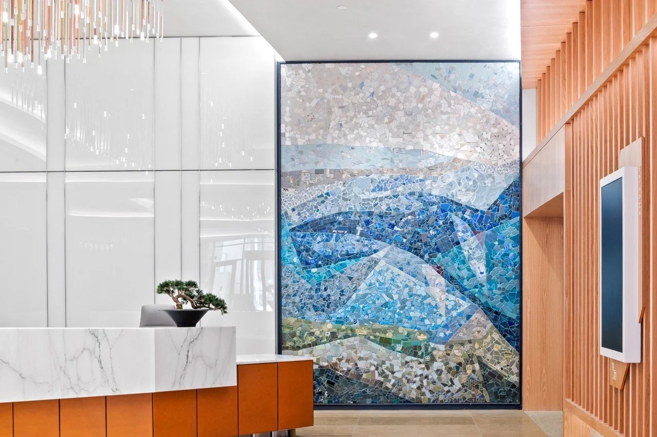 Interior of a modern lobby with a marble reception desk, a small bonsai tree, decorative wooden wall paneling, and a large abstract mosaic mural of a mountain landscape in shades of blue and gray.