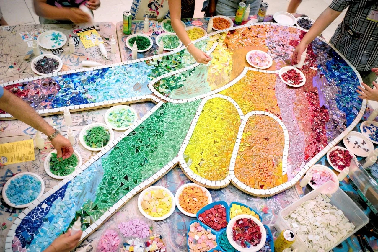 People creating a large mosaic art peace sign with a rainbow palette of mosaic tiles on a table.