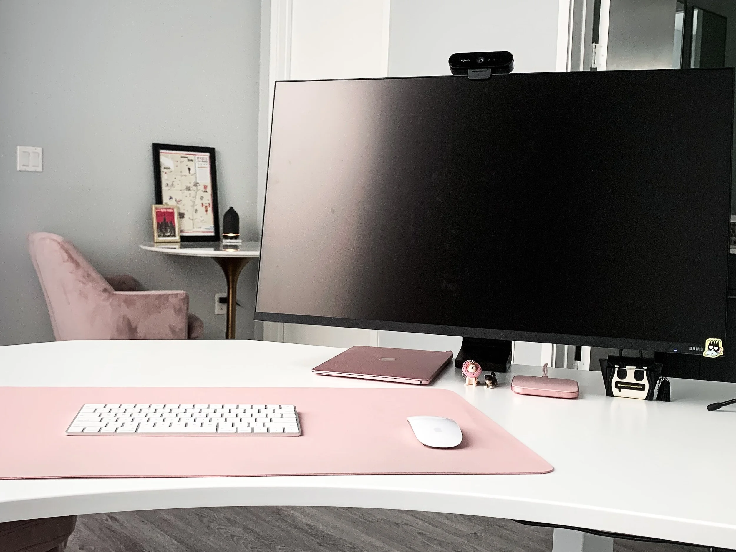 Modern white desk with a pink mouse pad, white keyboard, and a white mouse in the foreground. Behind the desk, a large black computer monitor with a webcam on top. Small pink and black accessories, including a pink laptop, a pink container, and decorative items. In the background, a pink armchair, a setup with framed pictures on a white table, and a gray wall with a light switch.
