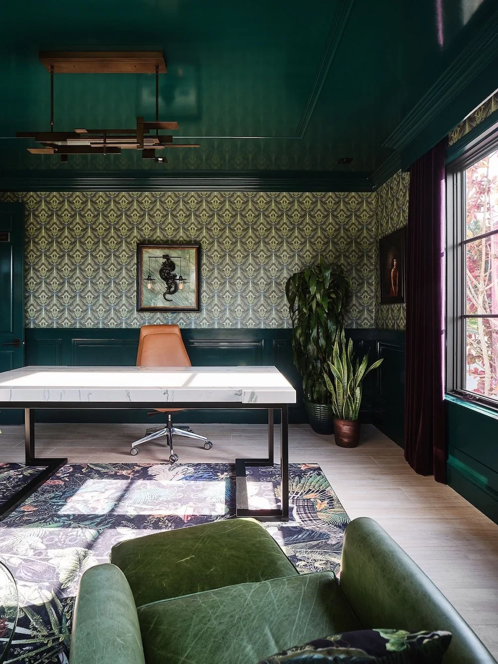 A modern office space featuring rich teal walls and ceiling, adorned with a patterned wallpaper. A large, minimalist white desk with marble accents sits primarily in the room, accompanied by a sleek, ergonomic chair with an orange hue. To the right,