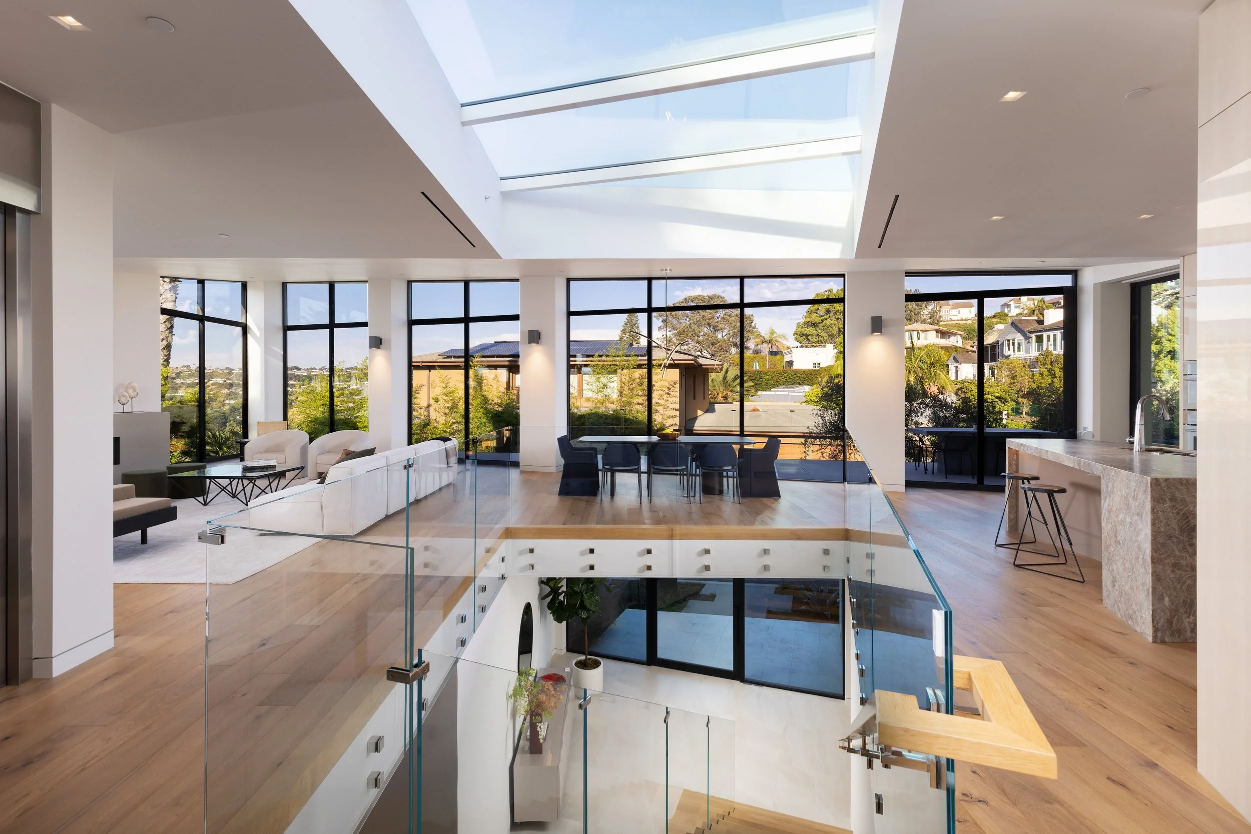 A modern, open-concept living space with large windows allowing natural light in. The room features a white sectional sofa and a black coffee table on a light rug, with sliding doors leading to an outdoor patio. In the background, there is a dining a