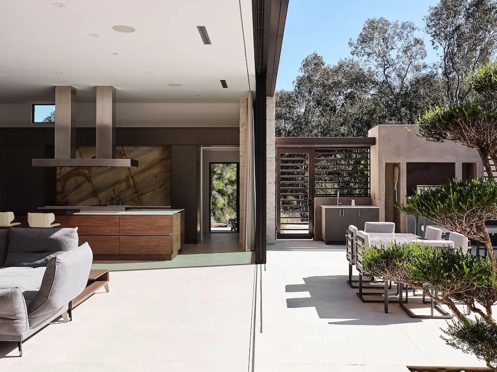A modern interior space featuring an open kitchen with dark cabinetry and a large central island. The kitchen is illuminated by natural light coming from large glass doors that open to an outdoor area. In the foreground, a grey sectional sofa is posi