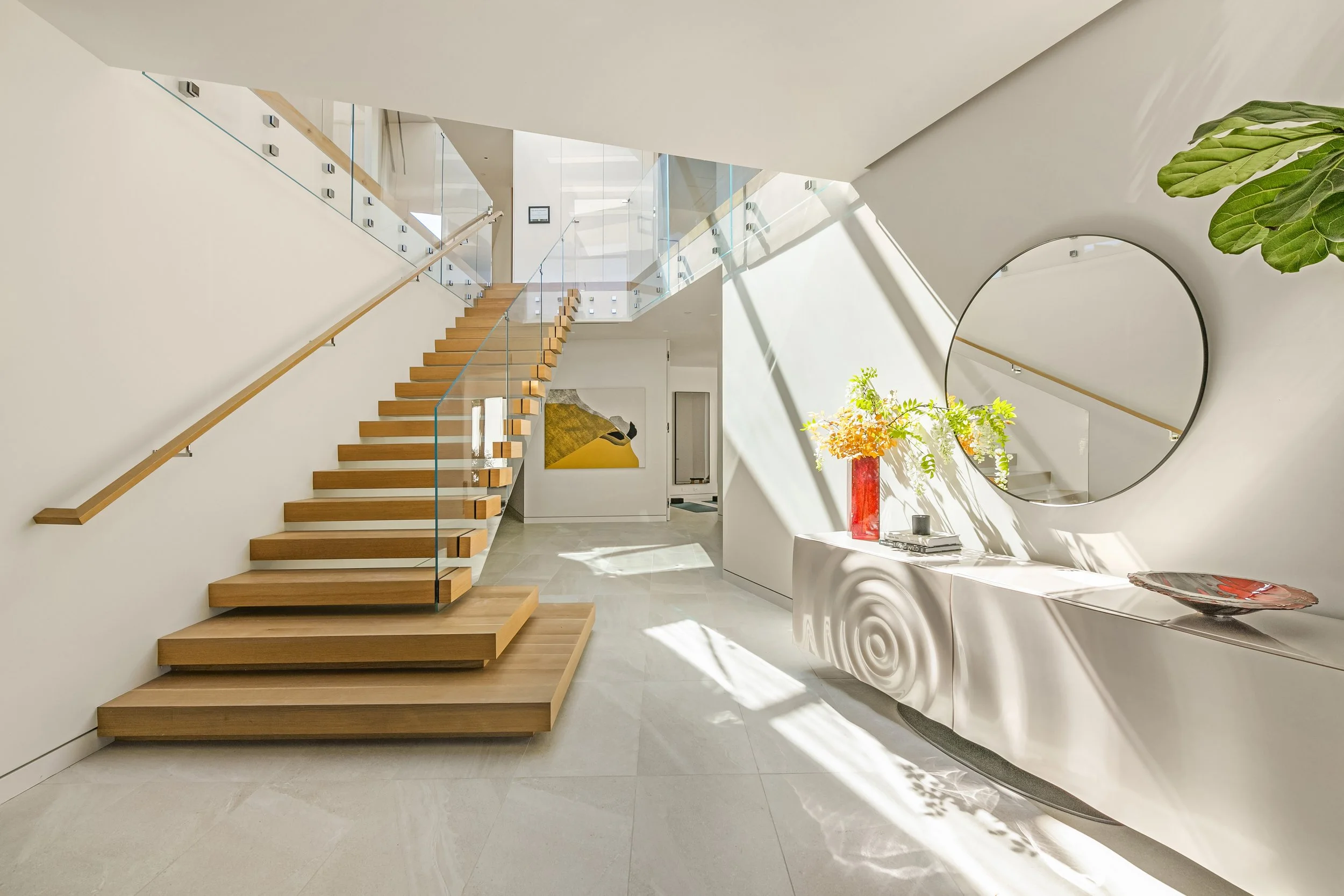 A modern, well-lit foyer featuring a wooden staircase with glass railings on the left side. To the right, a sleek, curved console table holds a red vase with flowers, a decorative plate, and a stack of books. A large round mirror is mounted on the wa