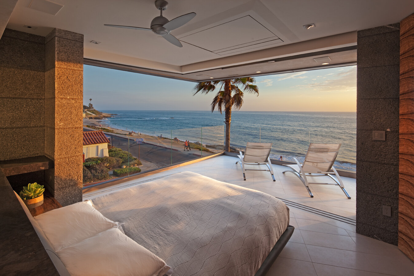 A luxurious bedroom featuring a large window with an ocean view. The room has a minimalist design with a neatly made bed, a bedside table, and a potted plant. Outside, two lounge chairs are positioned on a balcony, overlooking a beach and palm trees.