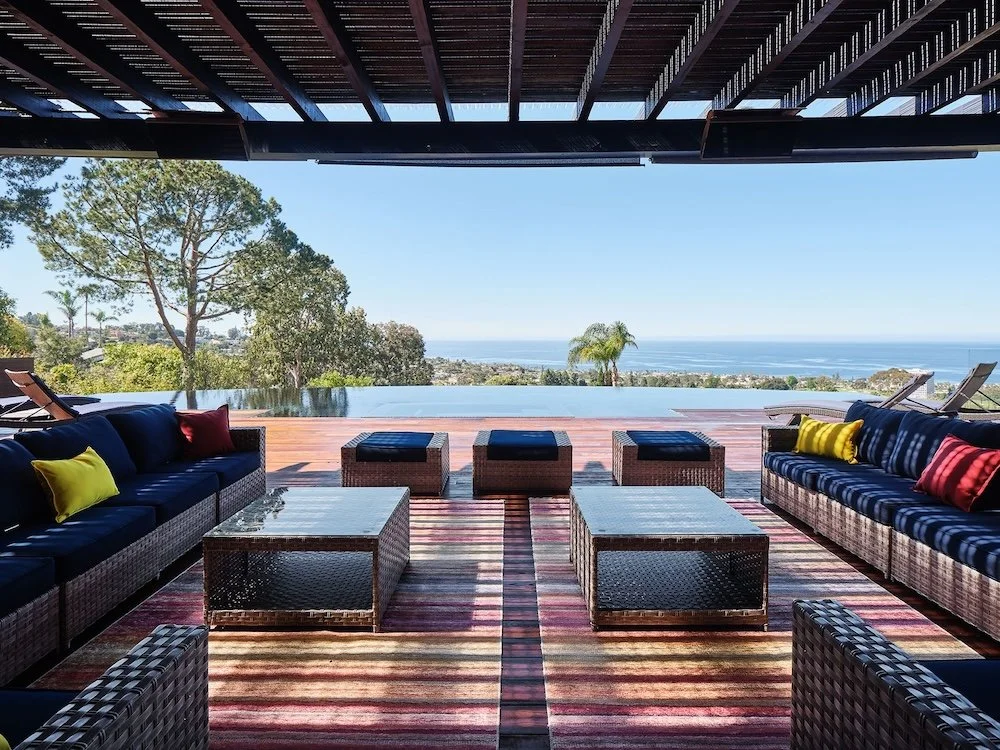 A spacious outdoor lounge area features a dark wicker sofa adorned with colorful cushions, a glass-topped coffee table, and matching ottomans on a vibrant striped rug. In the background, an infinity pool reflects the clear blue sky and overlooks a co