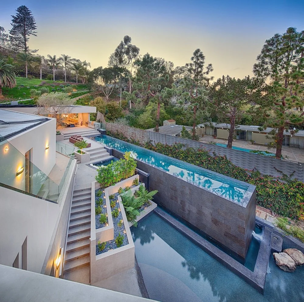 A modern, luxurious home is seen from an elevated angle, featuring a sleek design with large glass windows and a reflective swimming pool. The property includes tiered landscaping with lush greenery, palm trees, and colorful flowers. A long pool exte