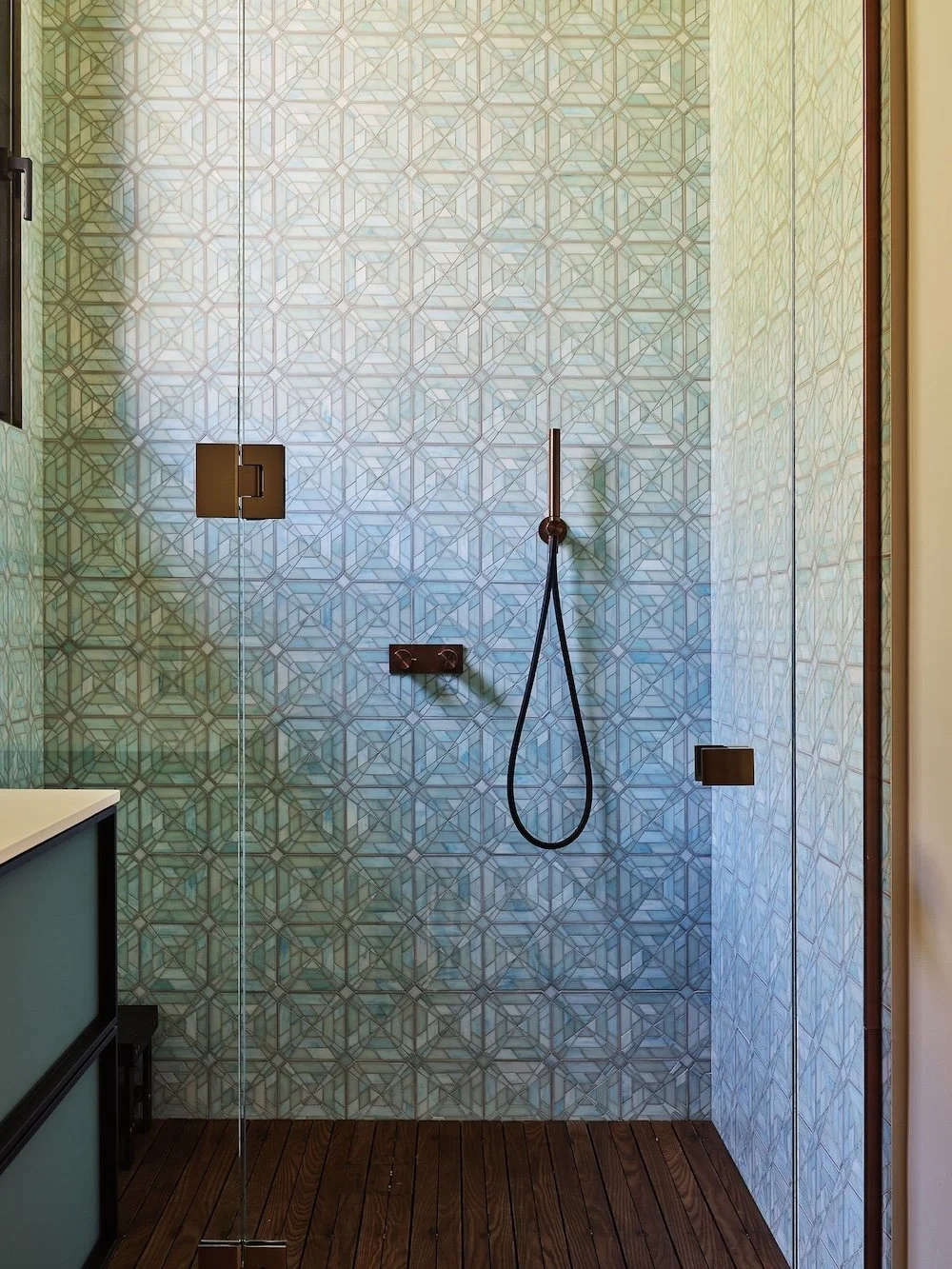 A modern shower area featuring a decorative, patterned wall in shades of blue and white, with geometric designs. The floor is made of wooden slats. A sleek, black showerhead hangs from the wall, and there are polished metal fixtures for control. A gl