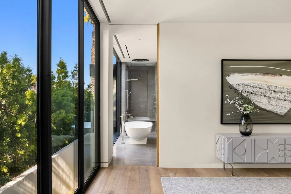 A bright, modern interior featuring large floor-to-ceiling windows that frame lush greenery outside. In the foreground, a stylish gray sideboard with decorative details holds a vase of flowers. Through an open doorway, a sleek bathroom is visible, sh