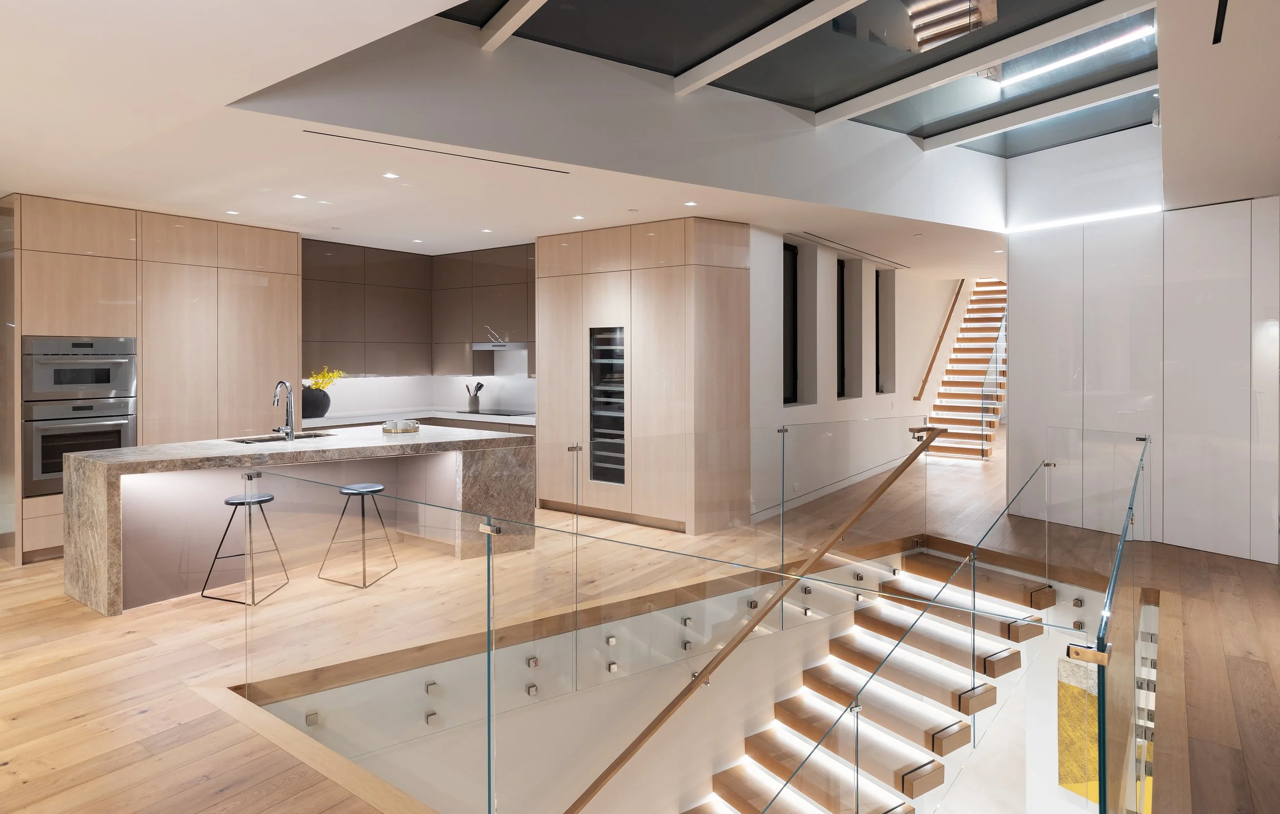 Modern kitchen and living area featuring sleek design elements. The space includes a large central island with a marble countertop, two black bar stools, and high-end appliances integrated into minimalist cabinetry. Large windows provide natural ligh