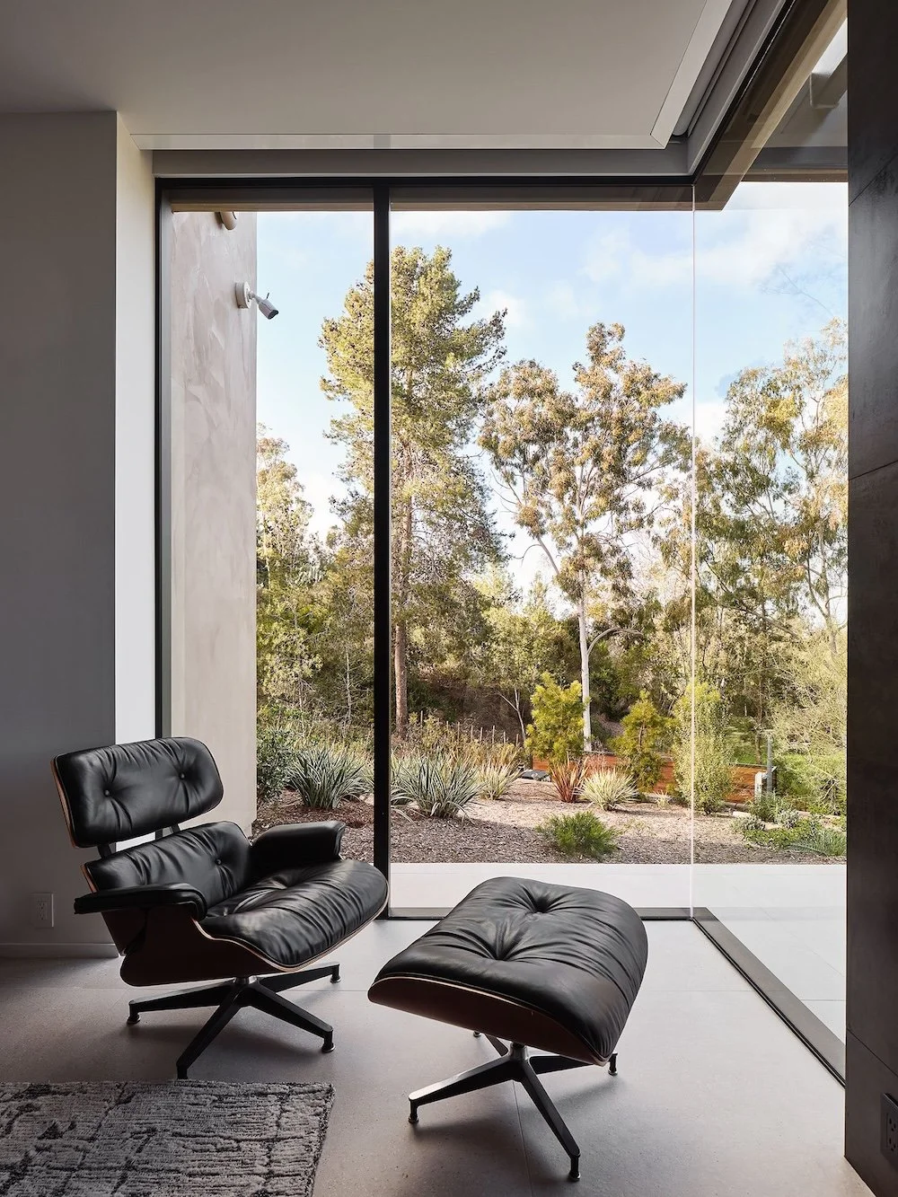 A modern lounge area featuring a sleek black leather chair and ottoman positioned by a large glass window. Outside, a landscaped garden with various trees and plants is visible under a partly cloudy sky. The interior has a neutral color palette with