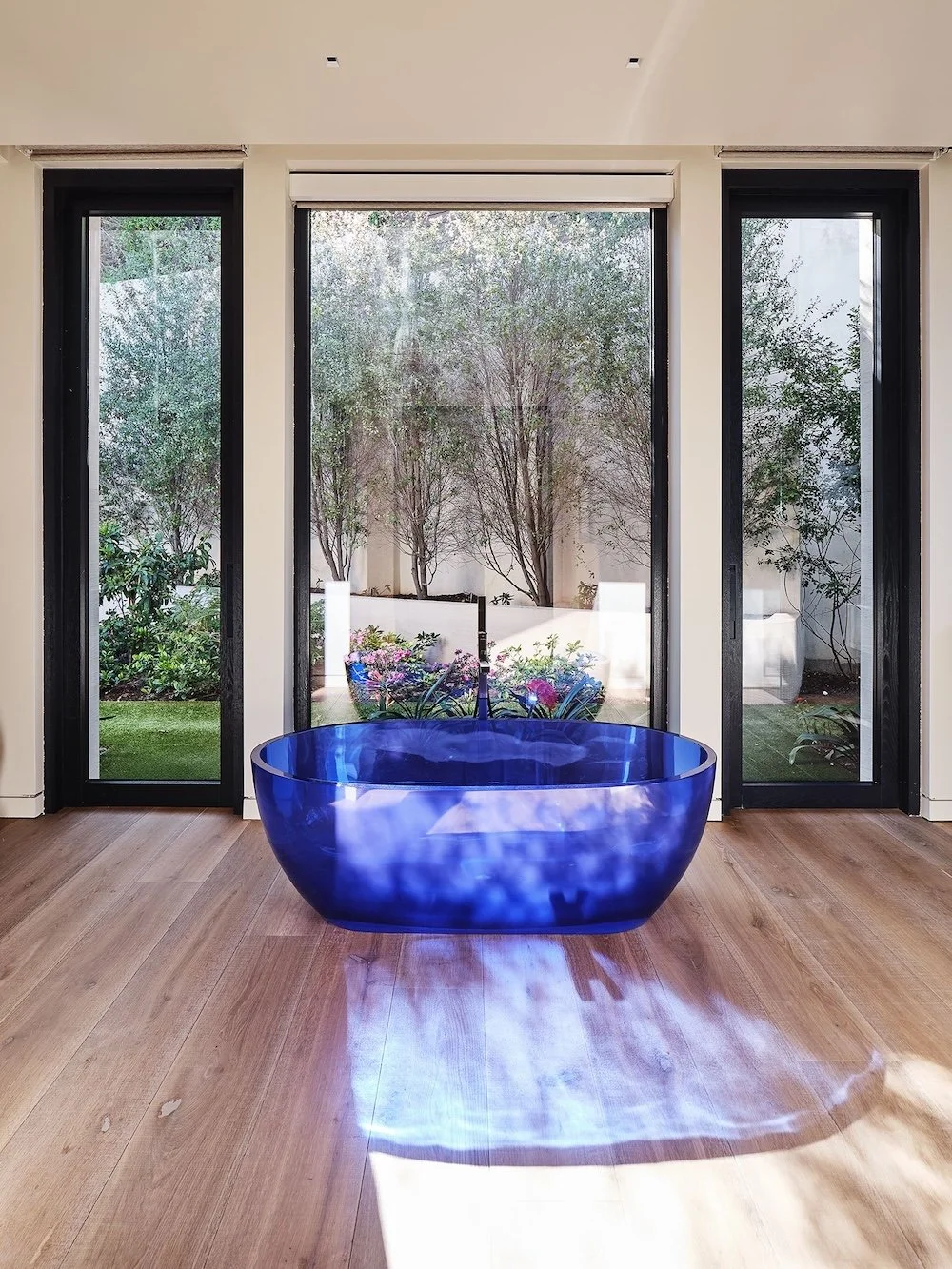 A modern interior space featuring a large, round, blue transparent bathtub in the center. Two tall black-framed windows on either side reveal a lush garden with greenery and flowering plants outside. The flooring is made of light-colored wood, and so
