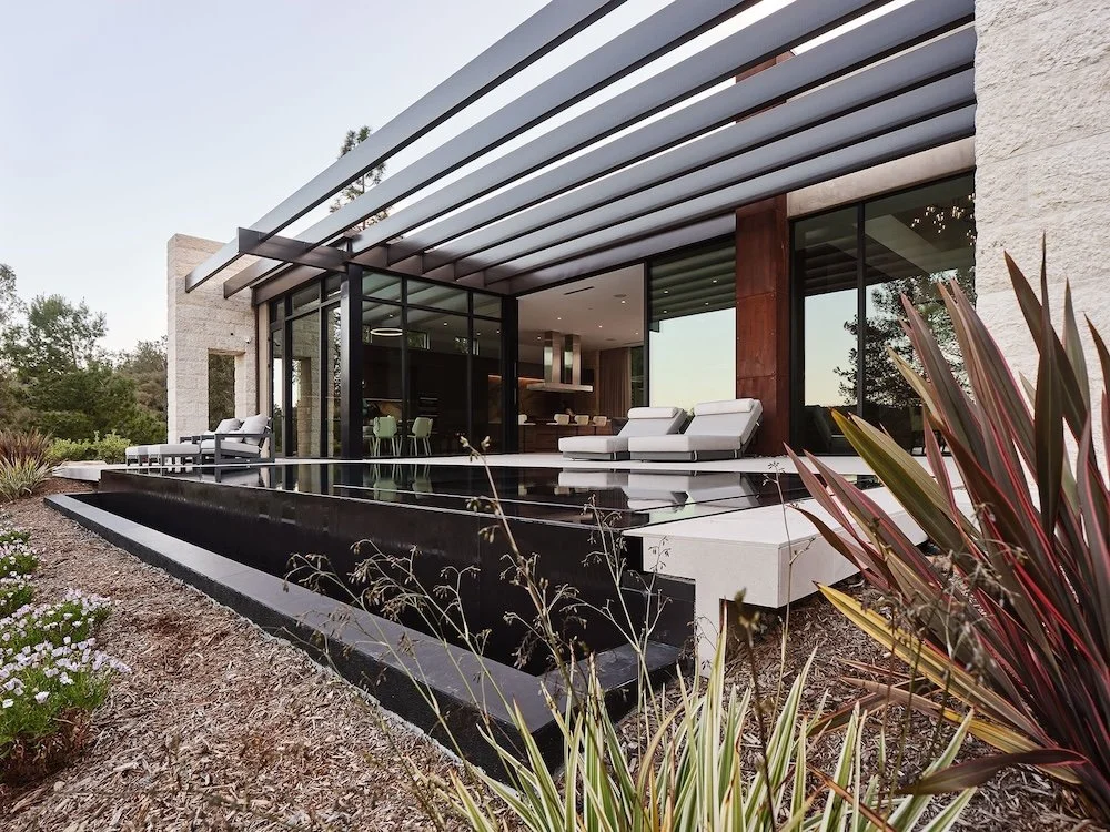 A modern house features a sleek design with large glass walls and a flat roof supported by horizontal beams. In front, there is a black reflecting pool surrounded by a landscaped area with various plants and shrubs. Two light gray lounge chairs are p