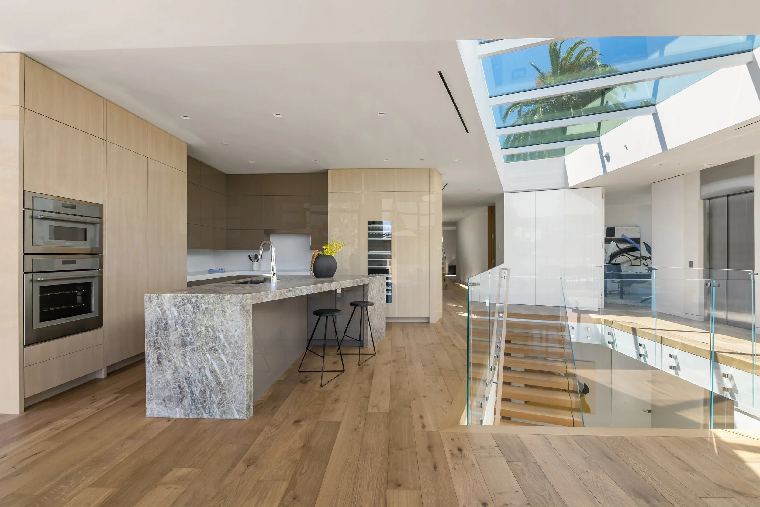 A modern kitchen featuring light wood cabinetry, sleek appliances, and a large gray stone island with two black stools. The space has large windows letting in natural light, revealing a staircase with glass railing in the background. The floor is mad