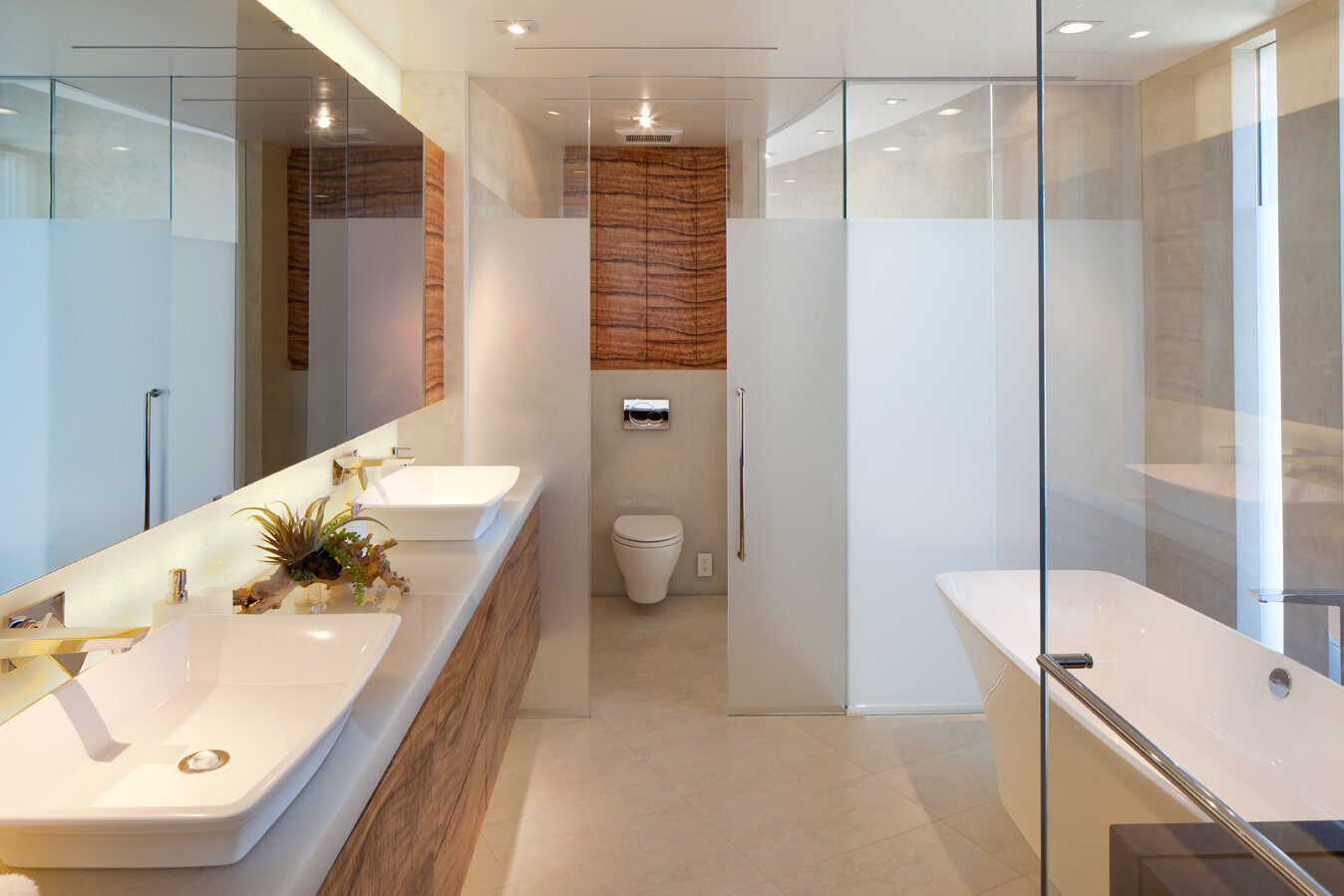 A modern bathroom featuring a double sink with sleek white basins atop a wooden vanity, accompanied by a decorative plant arrangement. The space is bright with natural light and illuminated by recessed lighting. To the right, a freestanding bathtub w