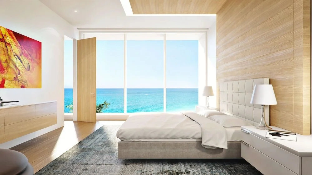 A modern bedroom featuring a large window that offers a view of the ocean. The walls are adorned with light wood and a colorful abstract painting in warm tones hangs on the wall. A plush bed with a beige headboard is positioned against the wall, and
