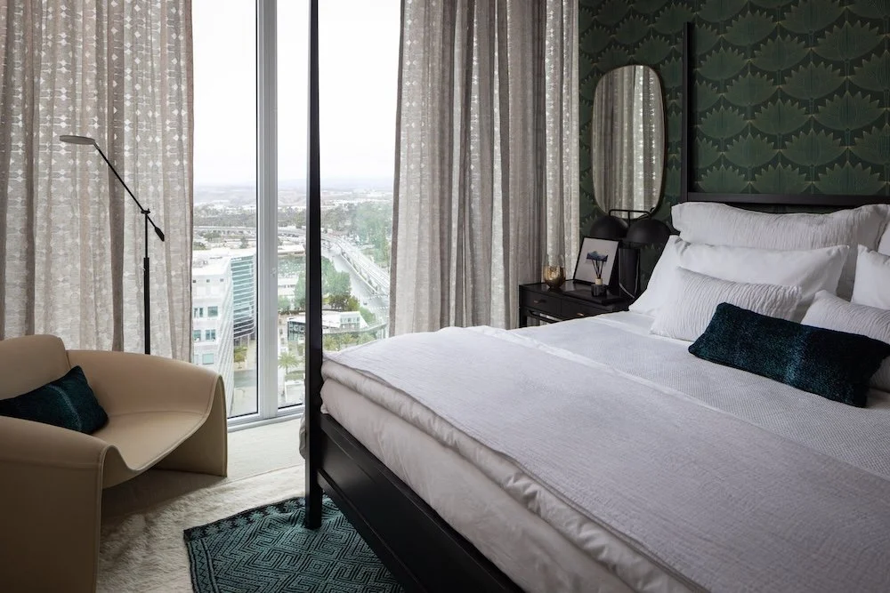 A stylish bedroom featuring a large window with sheer curtains, allowing natural light to fill the space. The room includes a black four-poster bed with neatly arranged white bedding and a decorative dark teal pillow. A modern beige accent chair is p