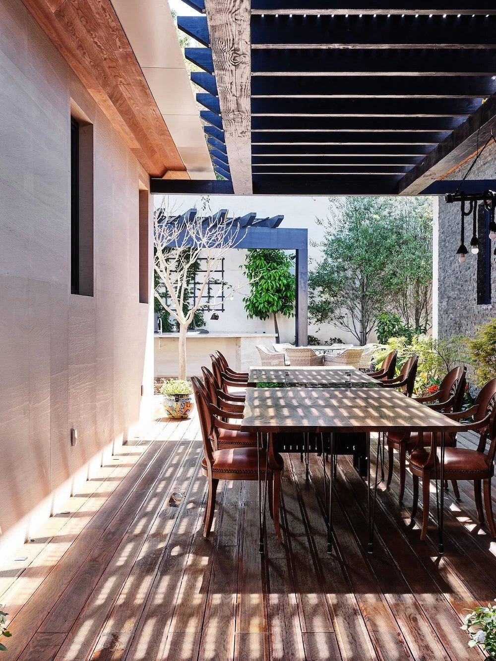 A serene outdoor dining area featuring a long, rectangular wooden table surrounded by several brown chairs. The setting is sheltered with a slatted pergola casting dappled shadows on the wooden deck. In the background, there are potted plants and sma