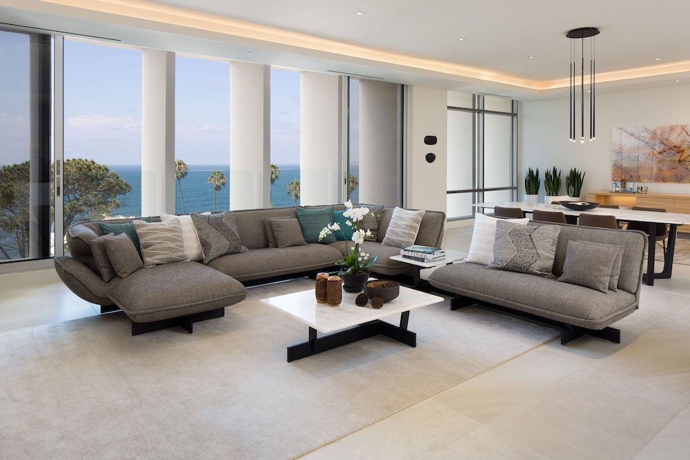 A modern living room with a large, grey sectional sofa adorned with various patterned cushions. In the foreground, a white coffee table holds a small black pot with white flowers and decorative items. The room features large windows with a view of th