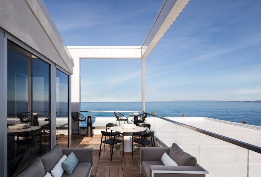 A modern outdoor terrace features a sleek, low couch with pillows and a round dining table surrounded by black chairs. The space is framed by large glass windows that reflect the blue sky. In the background, there is a stunning view of the ocean unde