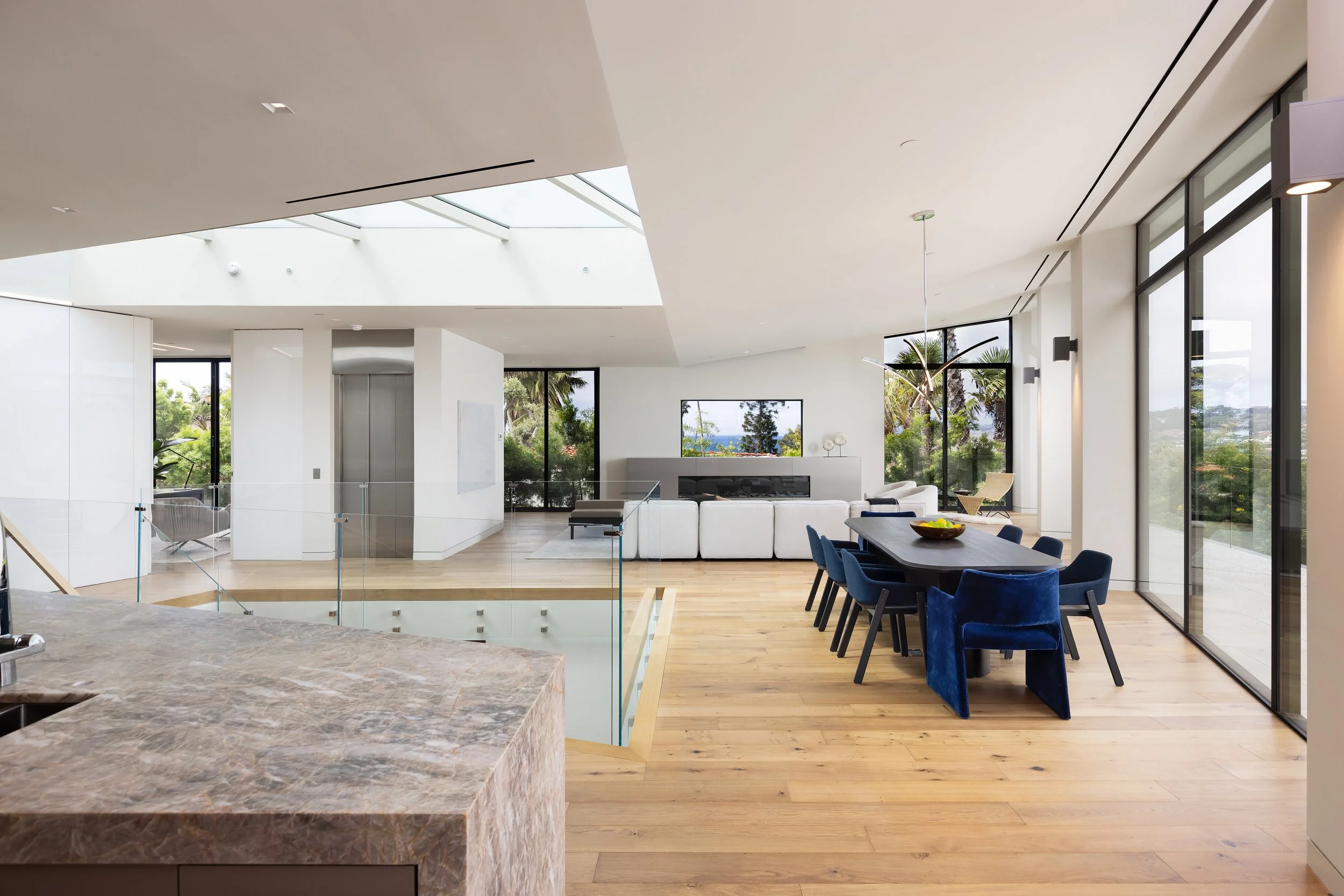 A spacious, modern living area featuring large windows that allow natural light to fill the room. The interior includes a kitchen with a sleek marble countertop, a dining area with a dark wooden table surrounded by blue upholstered chairs, and a comf