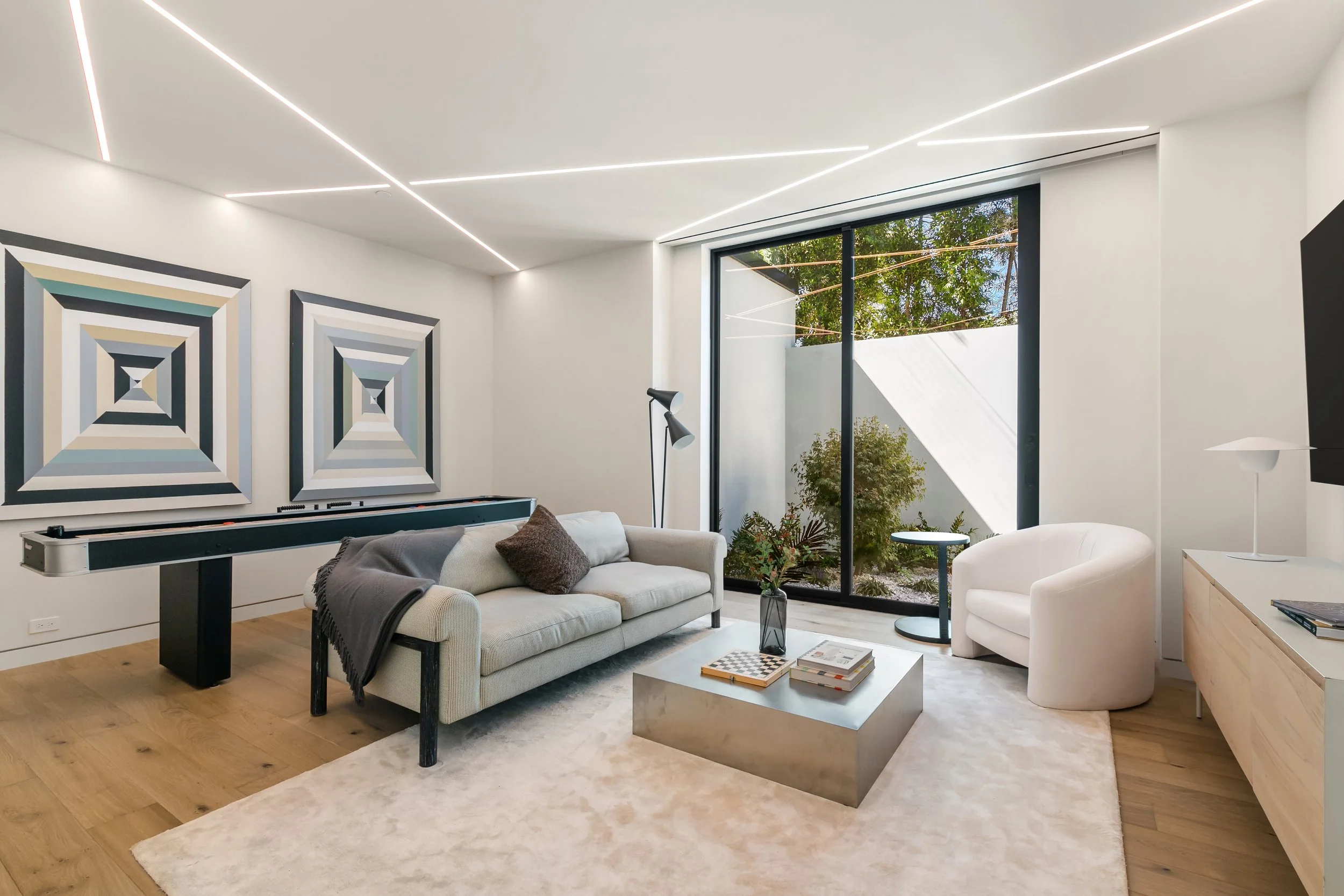 A modern living room featuring a light-colored sofa with a dark throw pillow and a square metallic coffee table. Two geometric artwork pieces are hung on the wall, and a black floor lamp stands beside the sofa. A large window offers a view of an outd