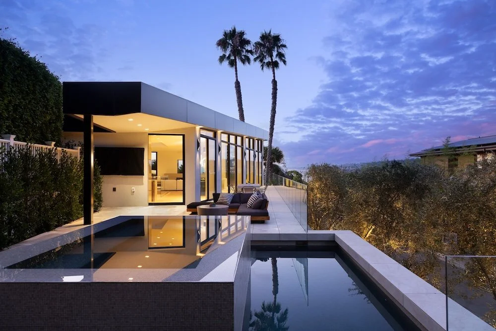 A modern house with large glass windows and a sleek black roof is set against a twilight sky filled with fluffy clouds. In the foreground, a tranquil swimming pool reflects the house and surrounding palm trees. A comfortable outdoor seating area with
