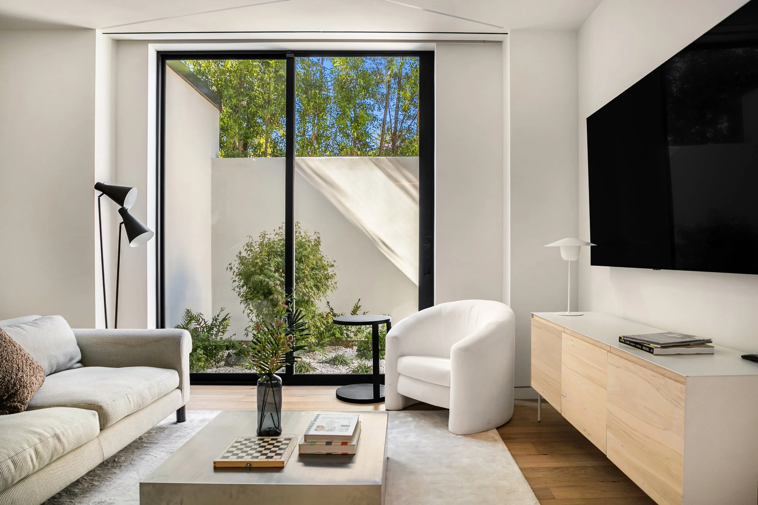 A modern living room featuring a light gray sofa with a decorative pillow, a white armchair, and a light wooden media console beneath a wall-mounted black TV. A small black side table sits next to the armchair. A large glass window allows natural lig