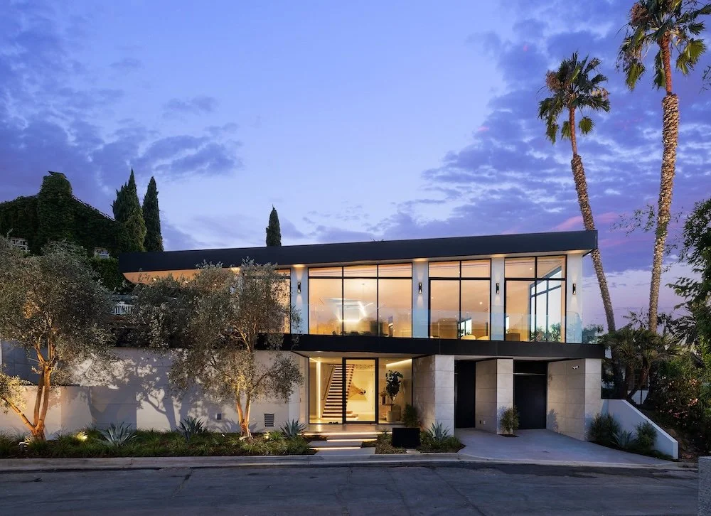 A modern two-story house with large glass windows and a sleek black roof is shown at dusk. The exterior features light-colored stone walls and is surrounded by landscaping, including olive trees and palm trees. Soft outdoor lighting illuminates the p