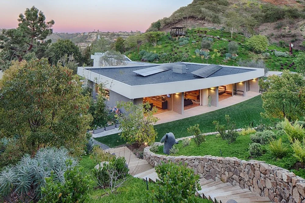 A modern single-story house with a flat roof featuring solar panels is set amidst lush greenery. The exterior walls are a light gray color, and large windows provide a view into the warmly lit interior. In the foreground, a stone pathway winds throug