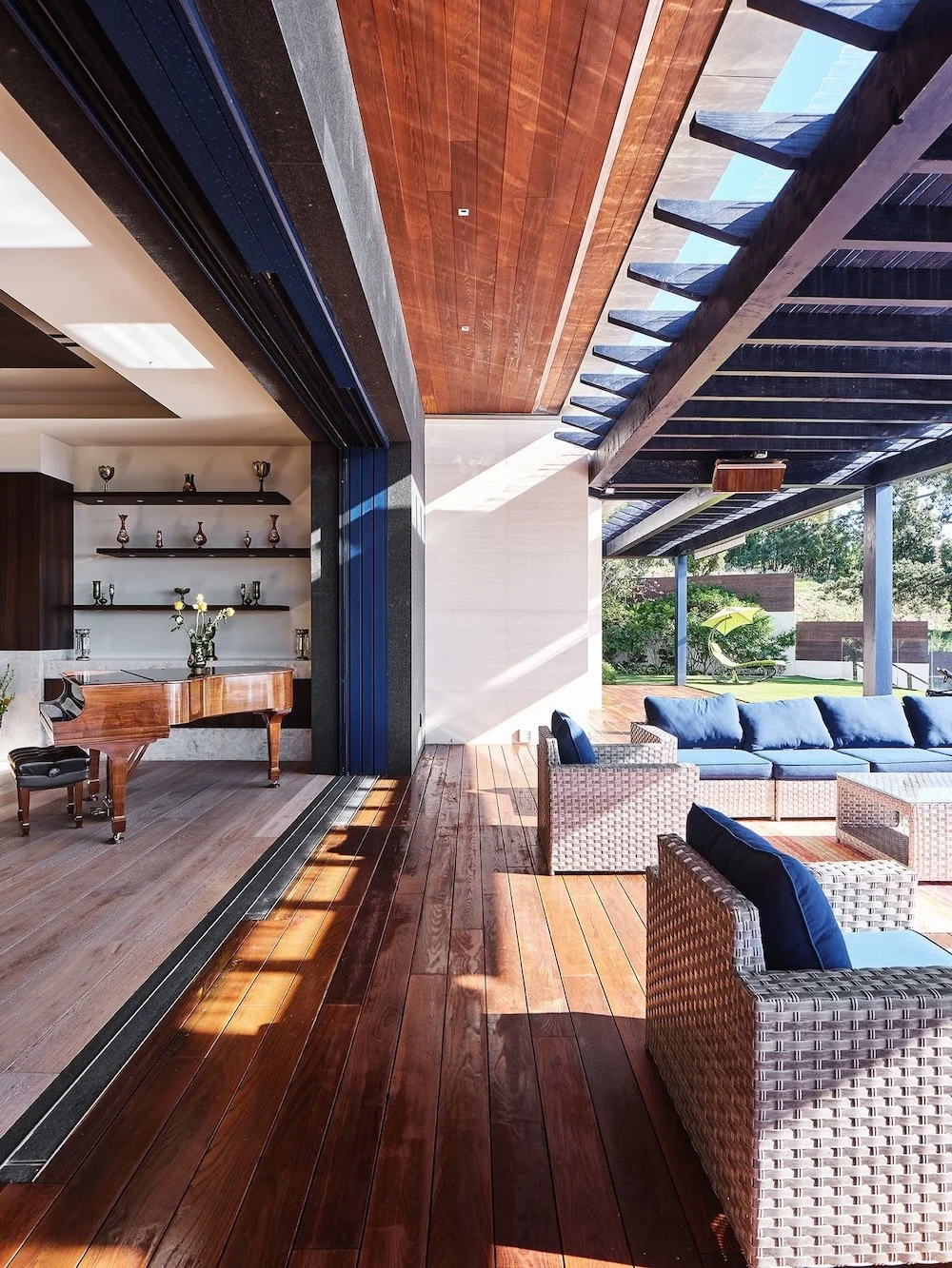 A spacious and modern outdoor living area with wooden flooring and open walls. To the left, a polished grand piano is set next to shelves displaying decorative vases. Comfortable seating consists of light-colored wicker chairs with blue cushions, arr