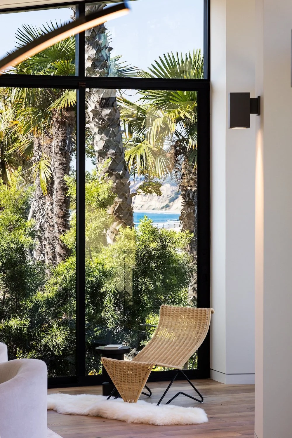A spacious, modern interior featuring a rattan chair on a soft white rug in front of a large window. Outside, palm trees and lush greenery are visible, along with a glimpse of the ocean in the background. The room has clean lines and natural light, c
