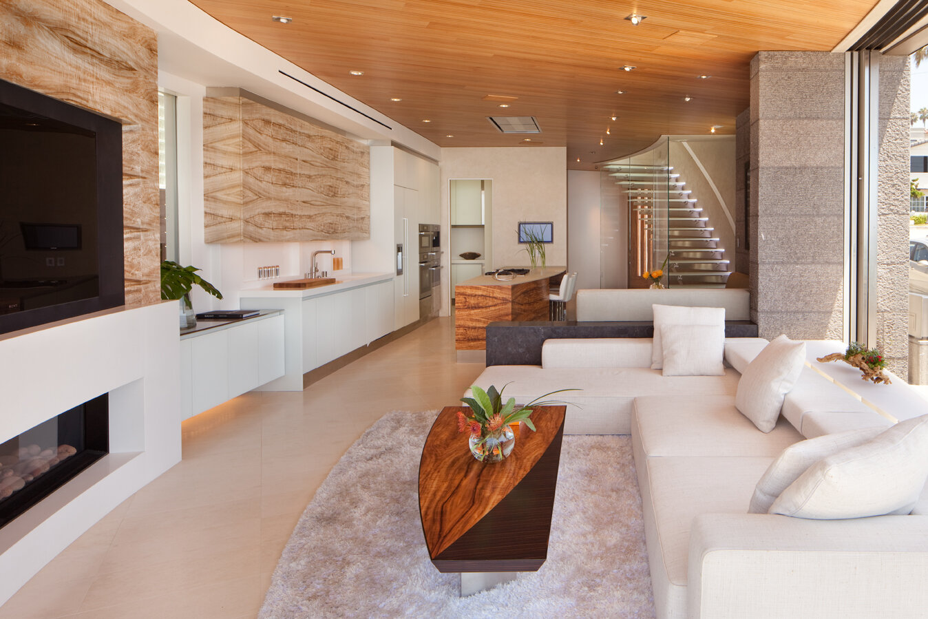 A modern, open-concept living space featuring a light-colored sofa with plush pillows, accompanied by a striking coffee table made of wood with a decorative plant. The room has wooden ceiling panels and walls adorned with natural stone. A sleek kitch