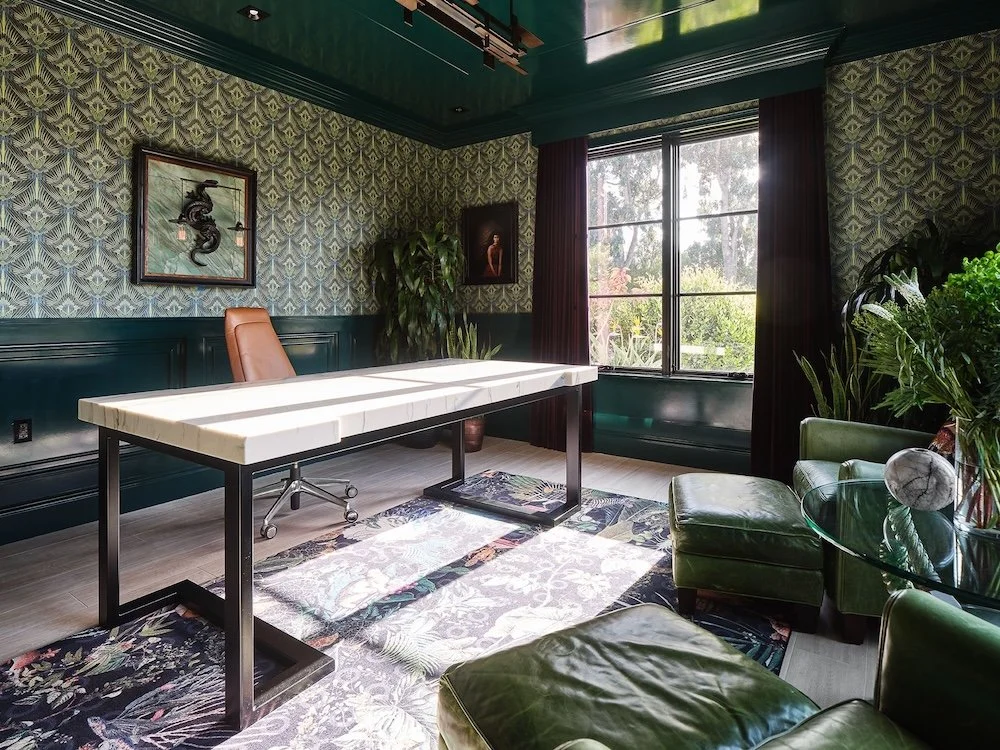 A modern office space with a dark green color scheme features a large white marble-topped desk with a black metal frame at the center. An ergonomic brown leather chair is positioned at the desk. The walls are adorned with patterned wallpaper, and art